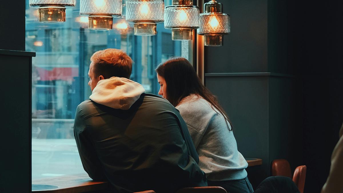 Two people sitting at a café table by a window under hanging lights, looking outside at a cityscape.