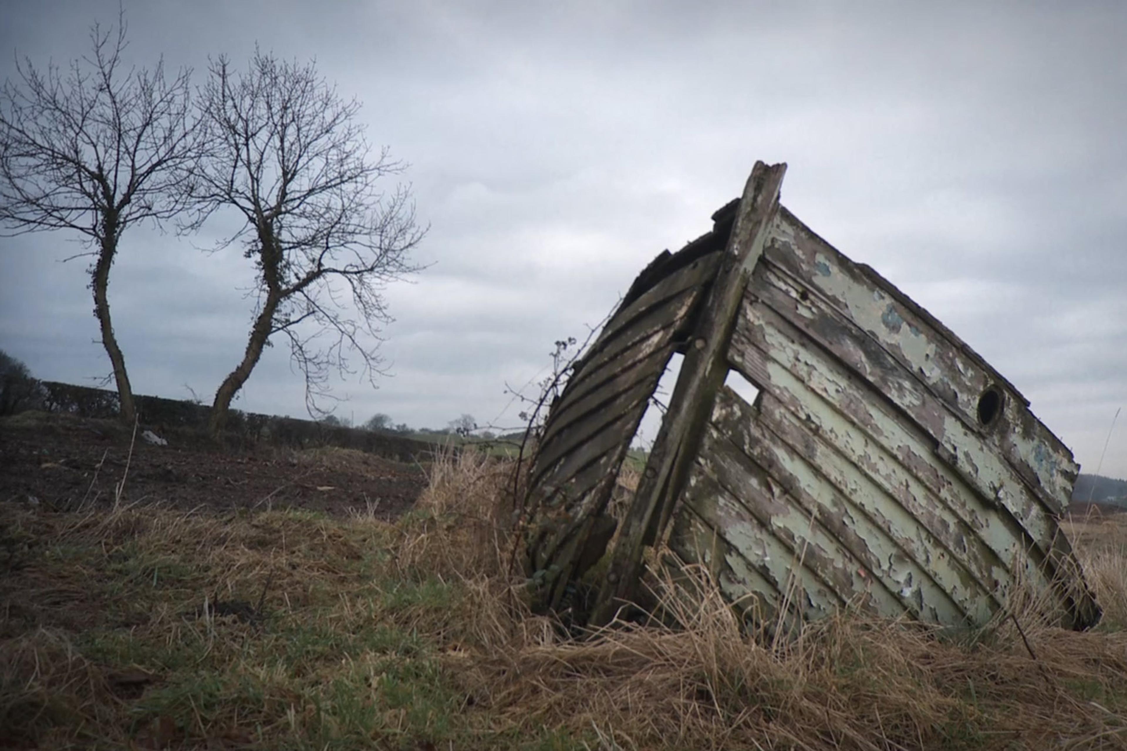 An old, weathered boat hull is grounded in a field with two bare trees under a cloudy sky.
