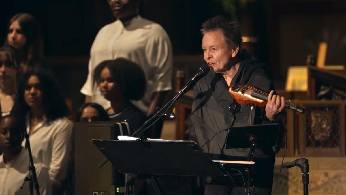 Photo of a person speaking into a microphone holding an electric string instrument with a choir in the background.