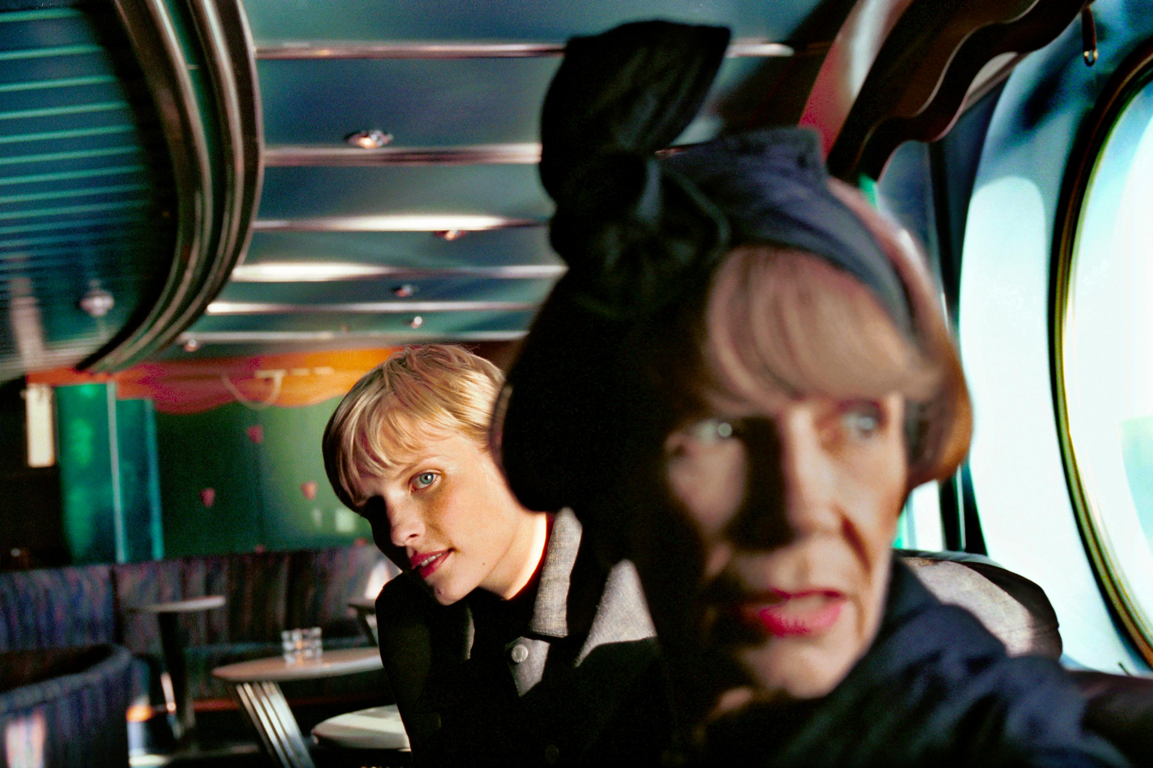 Photo of two women on a bus or train, one in focus looking back, the other blurred in the foreground, with teal interior tones.