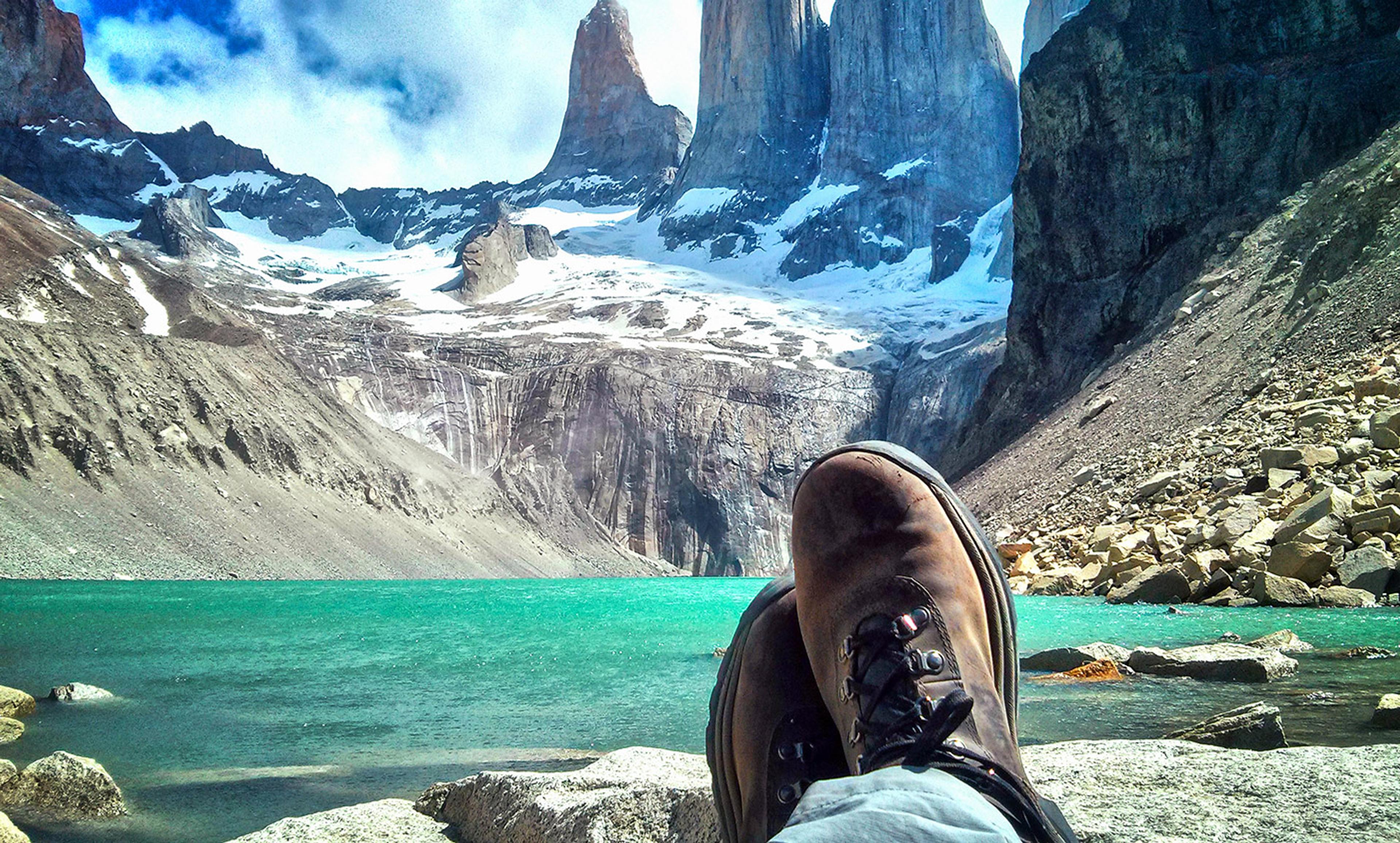 A person’s hiking boots resting on rocks by a turquoise lake with snowy mountains and cloudy blue sky in the background.