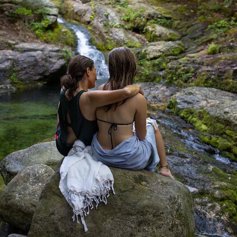 Two women wearing bathing suits sit on a rock by a waterfall in nature; one has her arm around the other. Towels are draped over the rock.