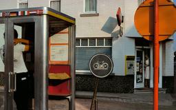 A man using a phone booth near traffic signs, including a bicycle sign, outside a building with a woman at the entrance.