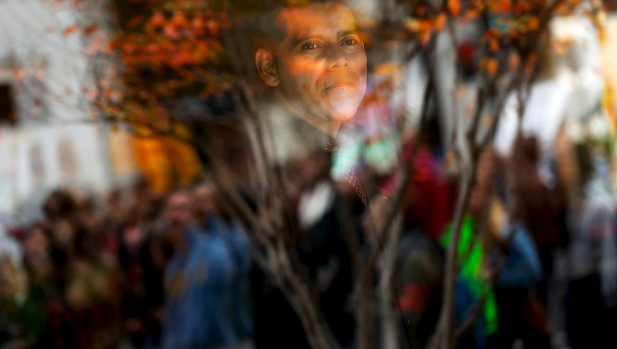 A man is seen through a windowpane with blurred people and autumn foliage shown in the reflection.