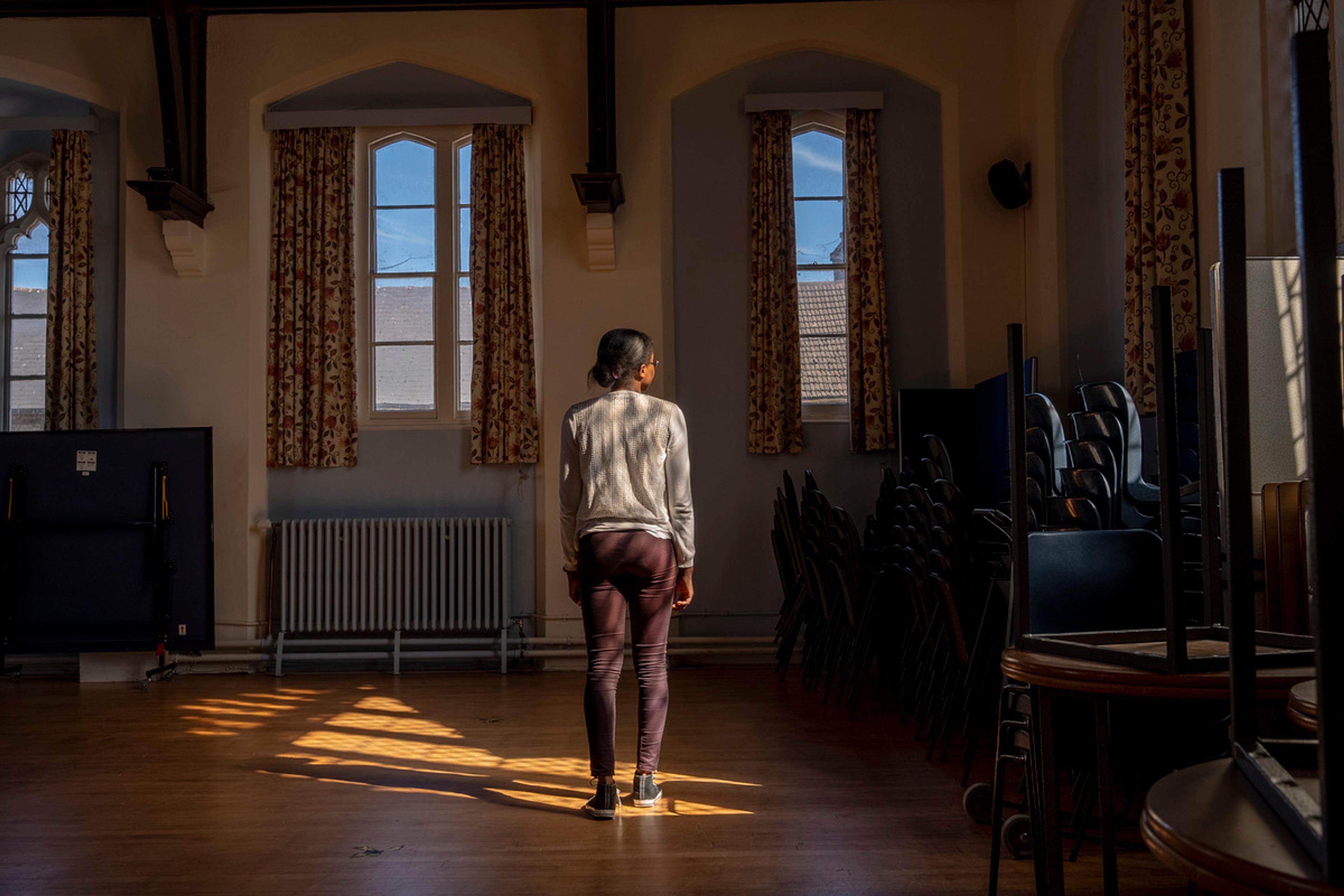 A woman stands in a patch of sunlight in a room with stacked chairs and windows framed by curtains.
