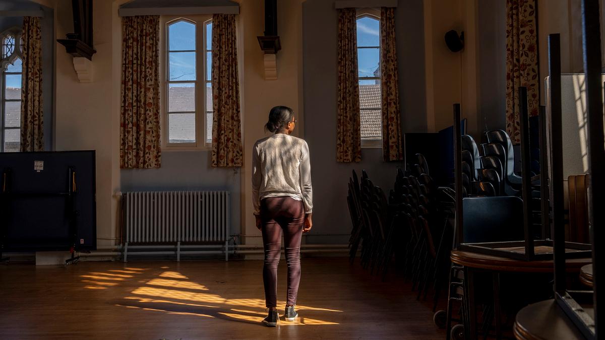 A woman stands in a patch of sunlight in a room with stacked chairs and windows framed by curtains.