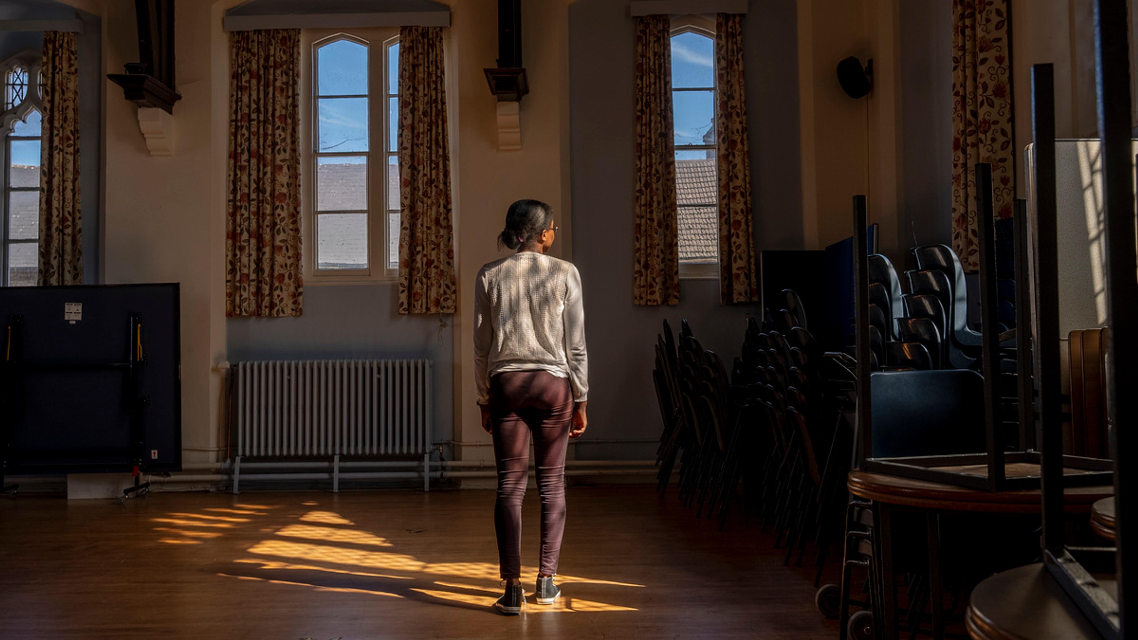 A woman stands in a patch of sunlight in a room with stacked chairs and windows framed by curtains.