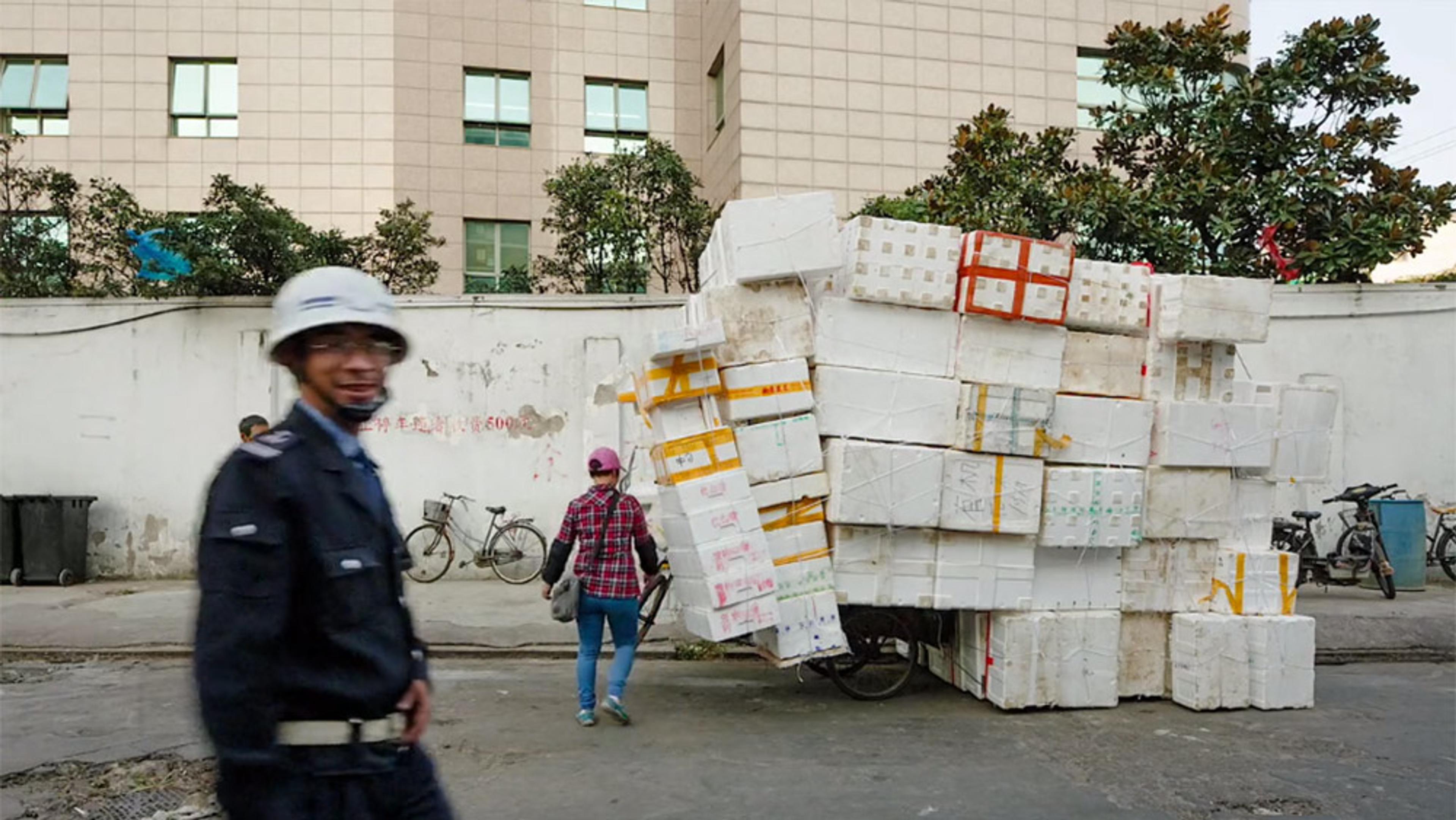 A smiling policeman walking past a heavily loaded tricycle with stacked boxes on an urban street, with buildings and bicycles in the background.