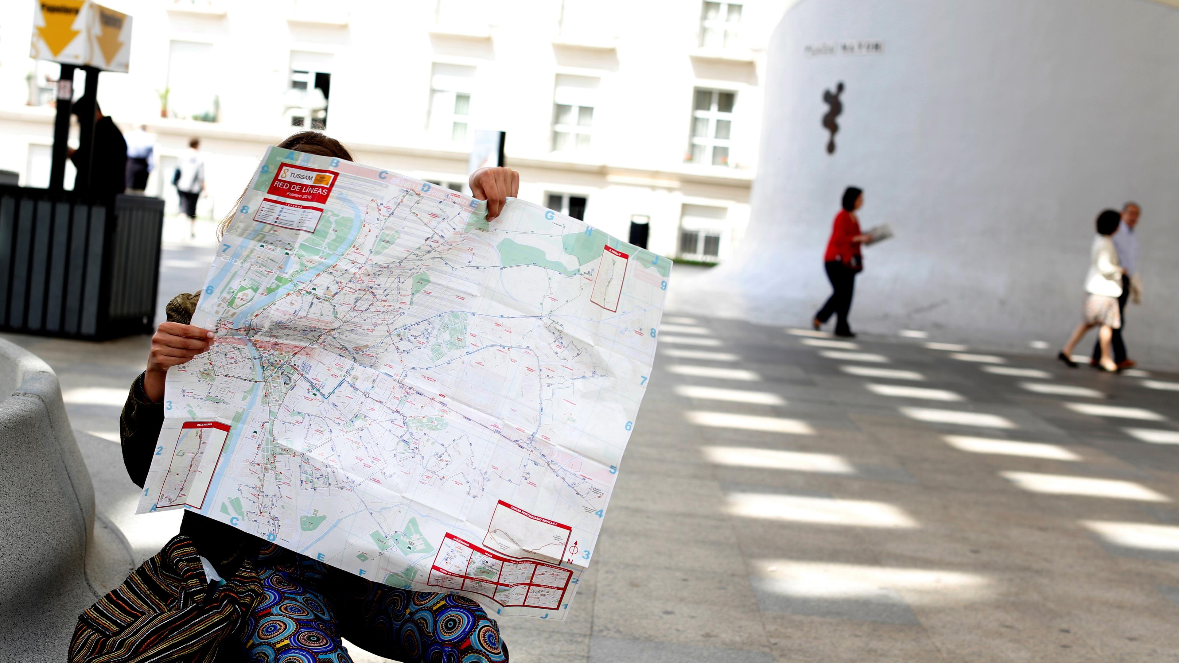 Photo of a person sitting and holding a large map covering their face outdoors with people walking in the background.