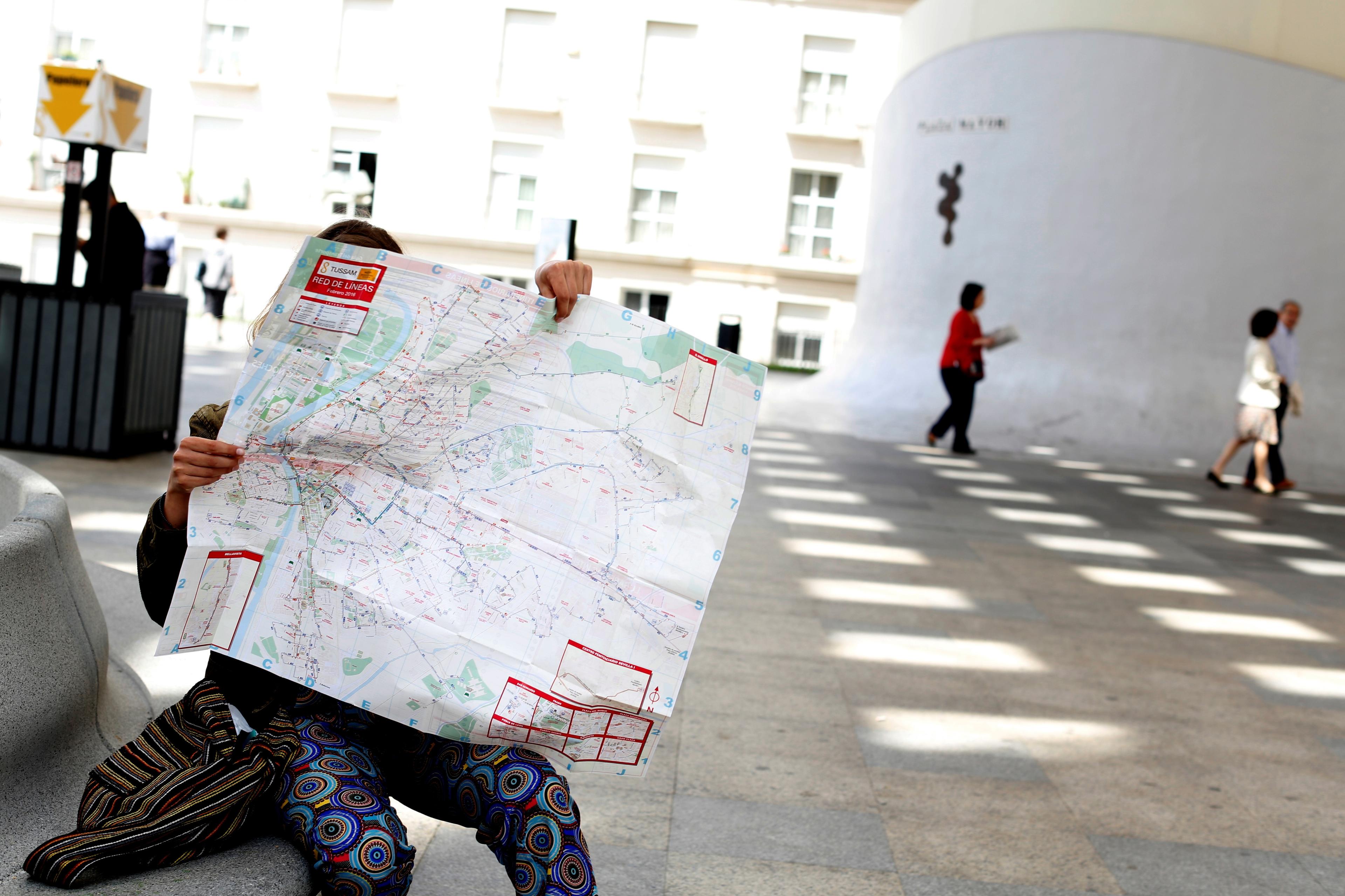 Photo of a person sitting and holding a large map covering their face outdoors with people walking in the background.
