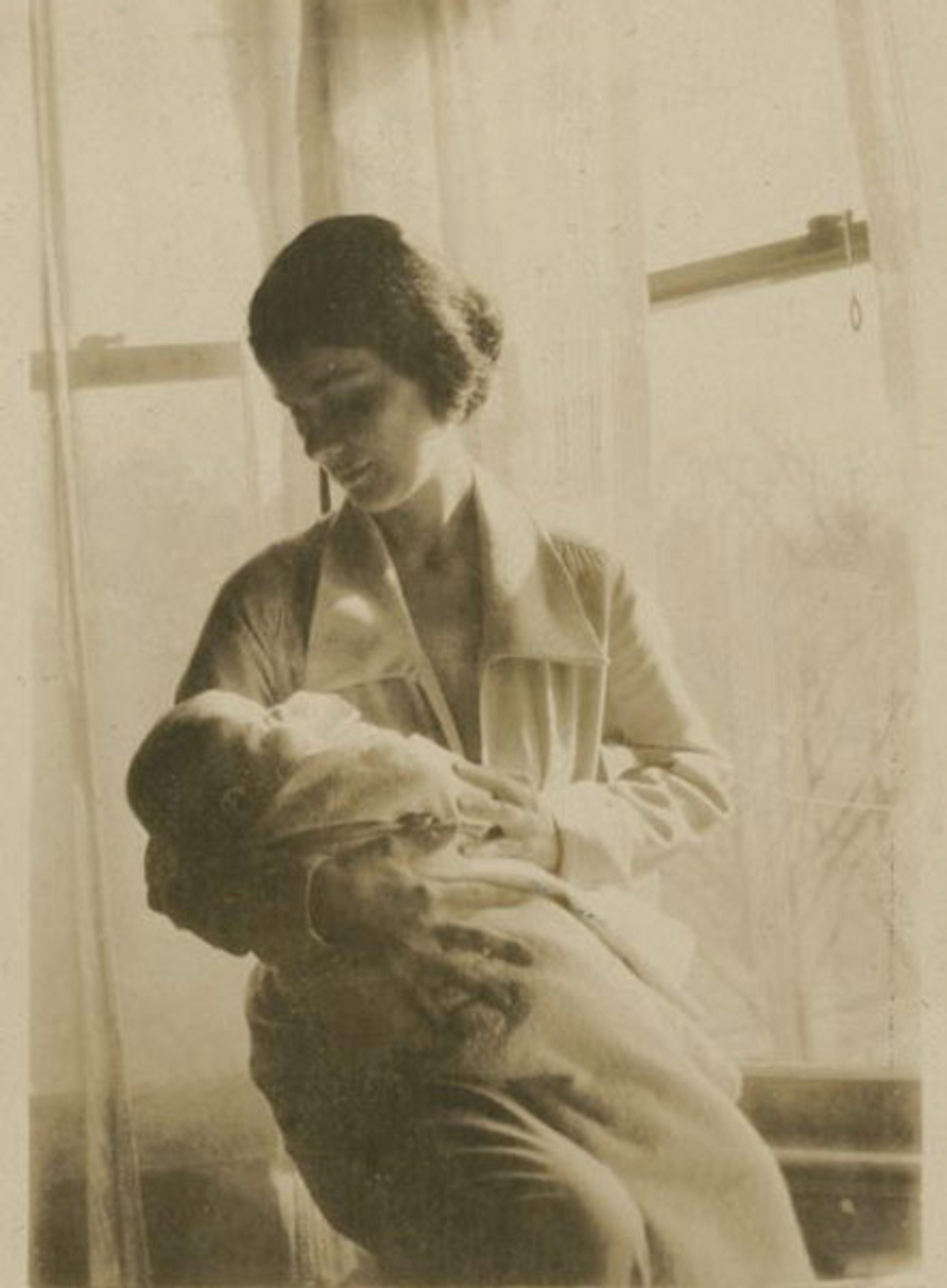 Sepia-toned photo of a woman sitting by a window holding a baby, softly lit by natural light through sheer curtains.