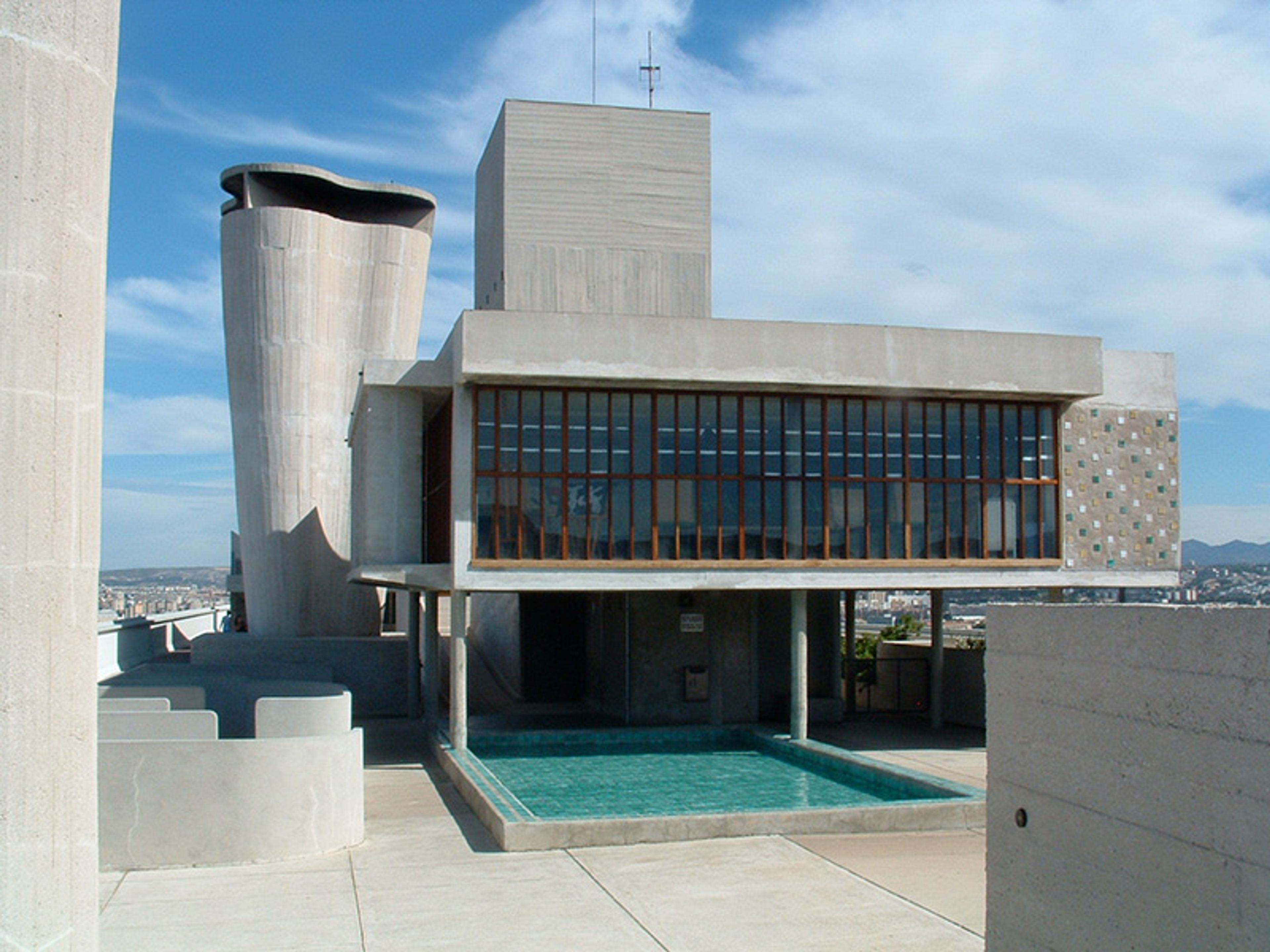 A modernist concrete building with large windows, a distinctive tall cylindrical structure, and a small pool in the foreground.