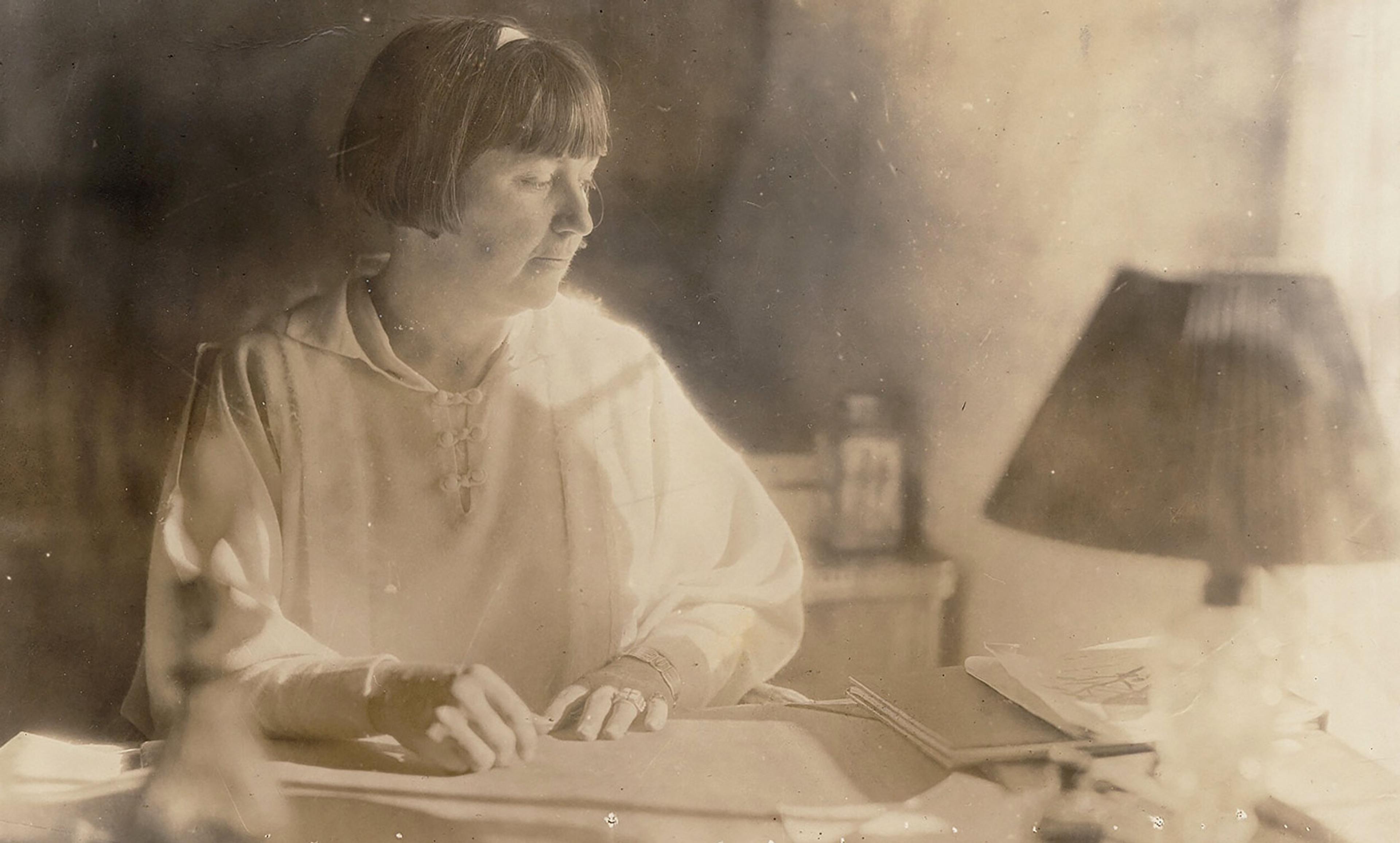 Vintage photo of a woman with a bob haircut, seated at a desk with papers and a lamp, looking down while writing or reading.