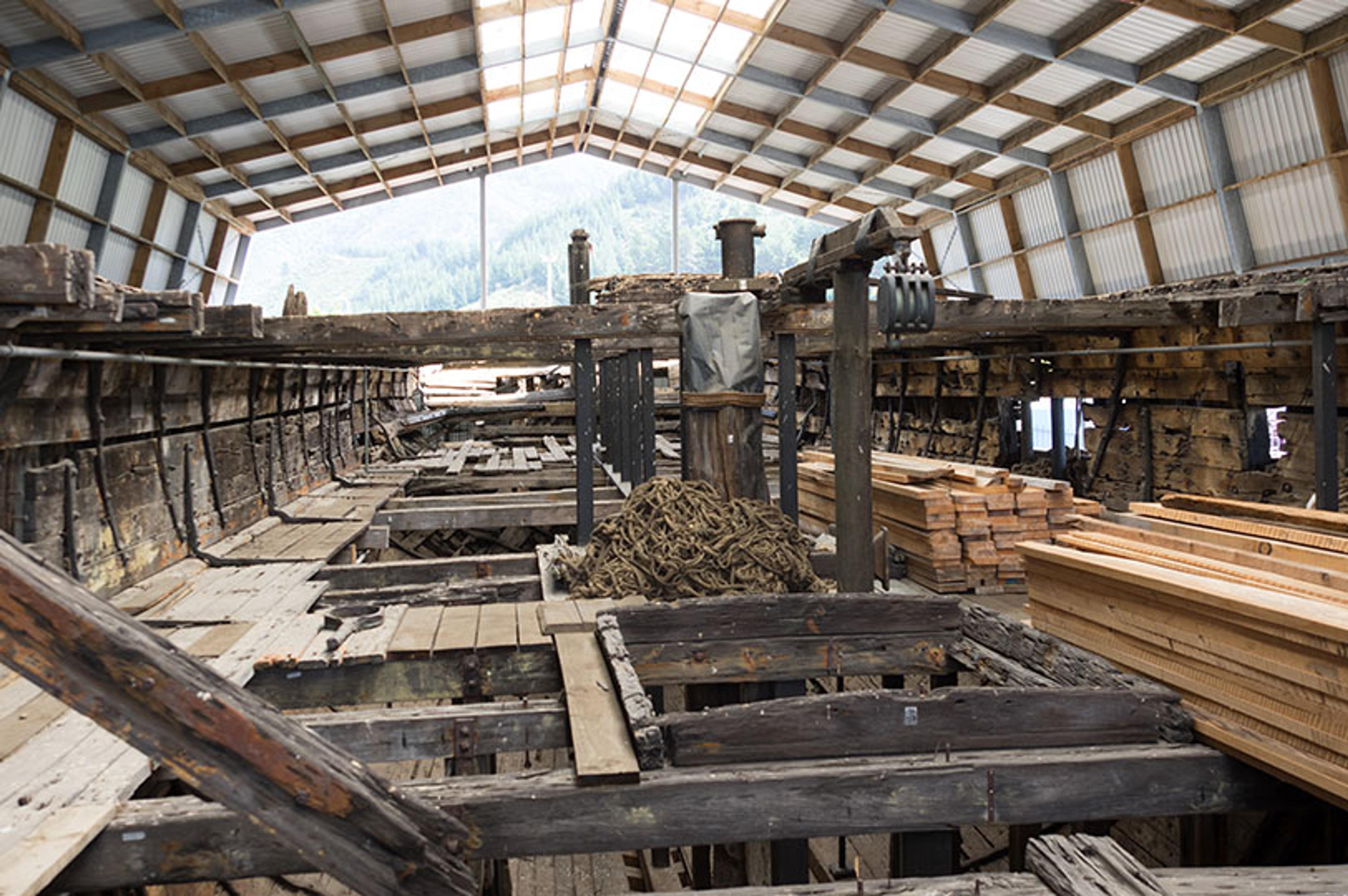 The interior of a large, partially restored wooden ship under a metal roof, with piles of lumber and coiled ropes.