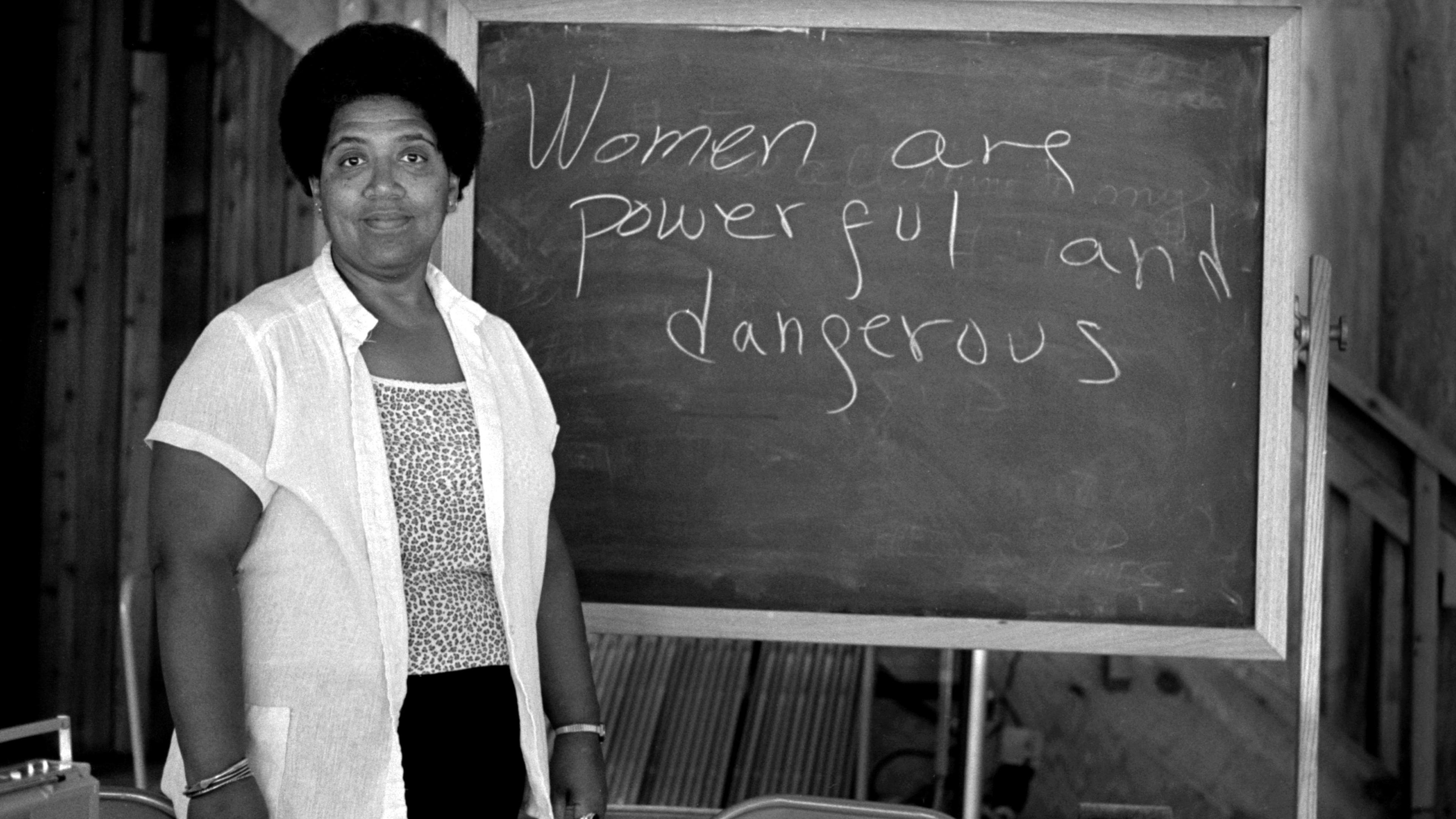 Black and white photo of a person by a chalkboard with “Women are powerful and dangerous” written on it.