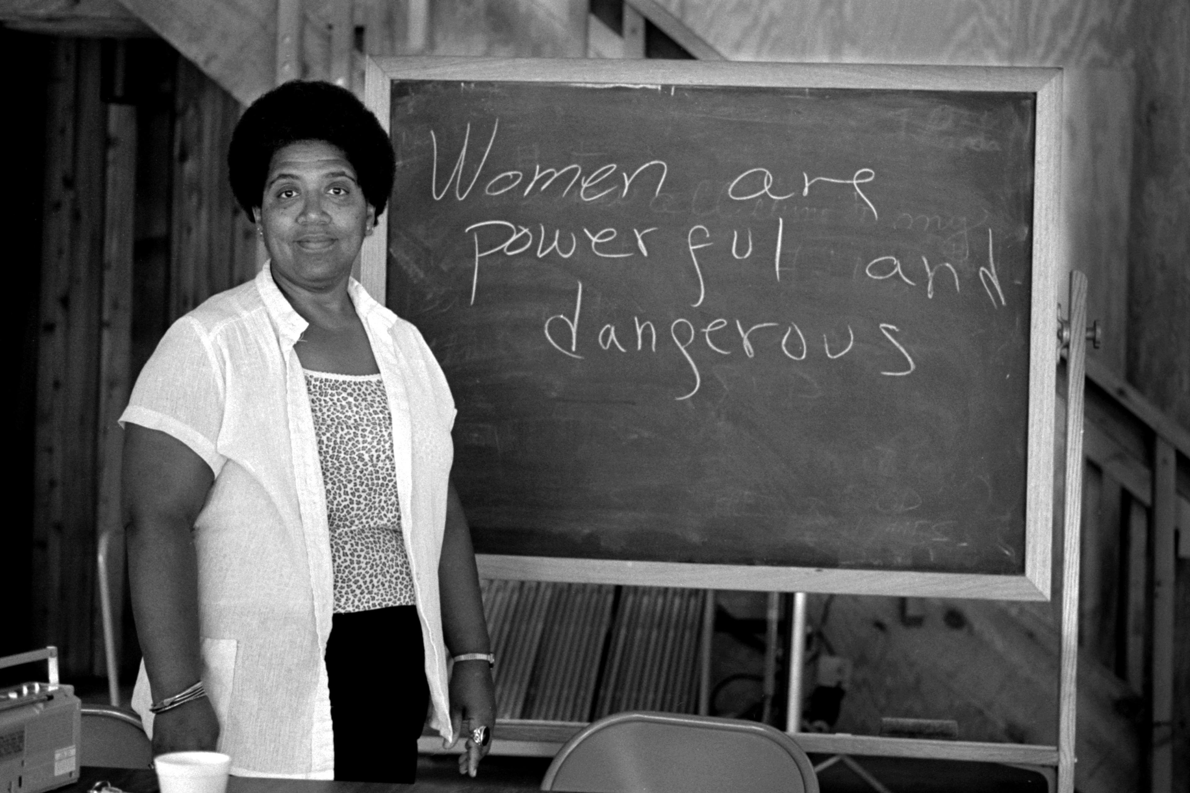 Black and white photo of a person by a chalkboard with “Women are powerful and dangerous” written on it.