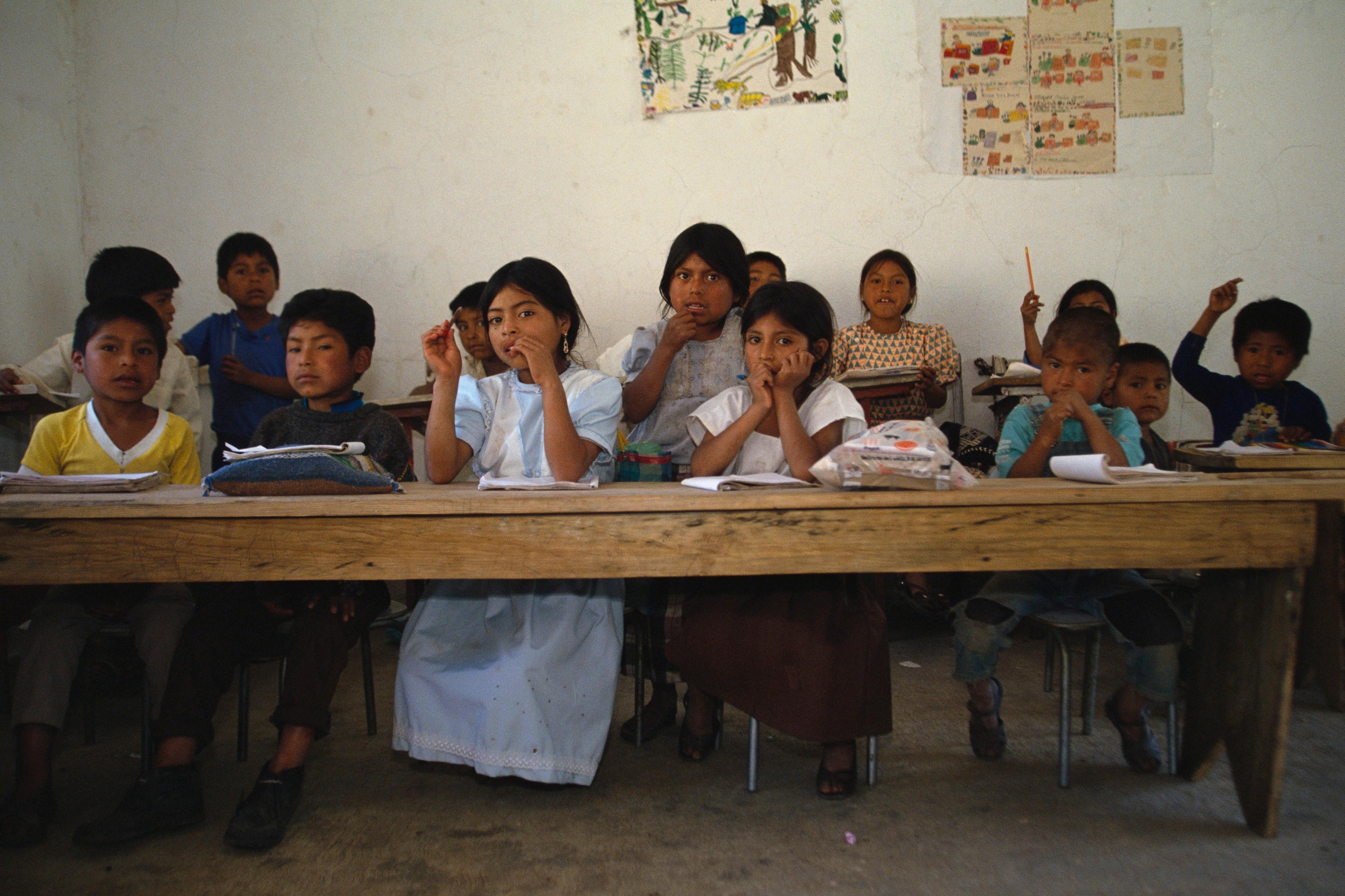 Photo of children sitting at a wooden desk in a classroom with drawings on the wall.