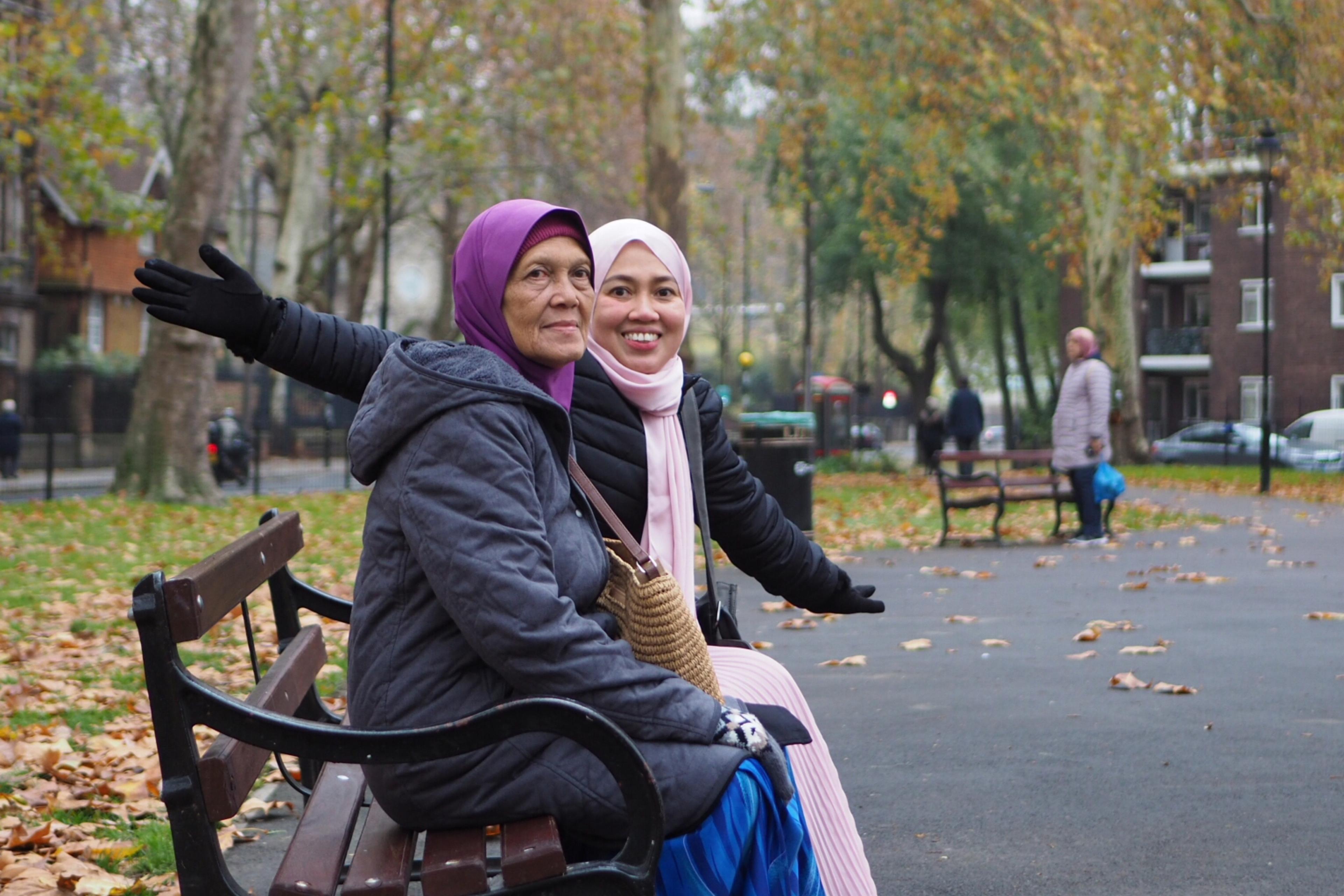 Photo of two women sitting on a park bench in autumn, surrounded by fallen leaves and trees.