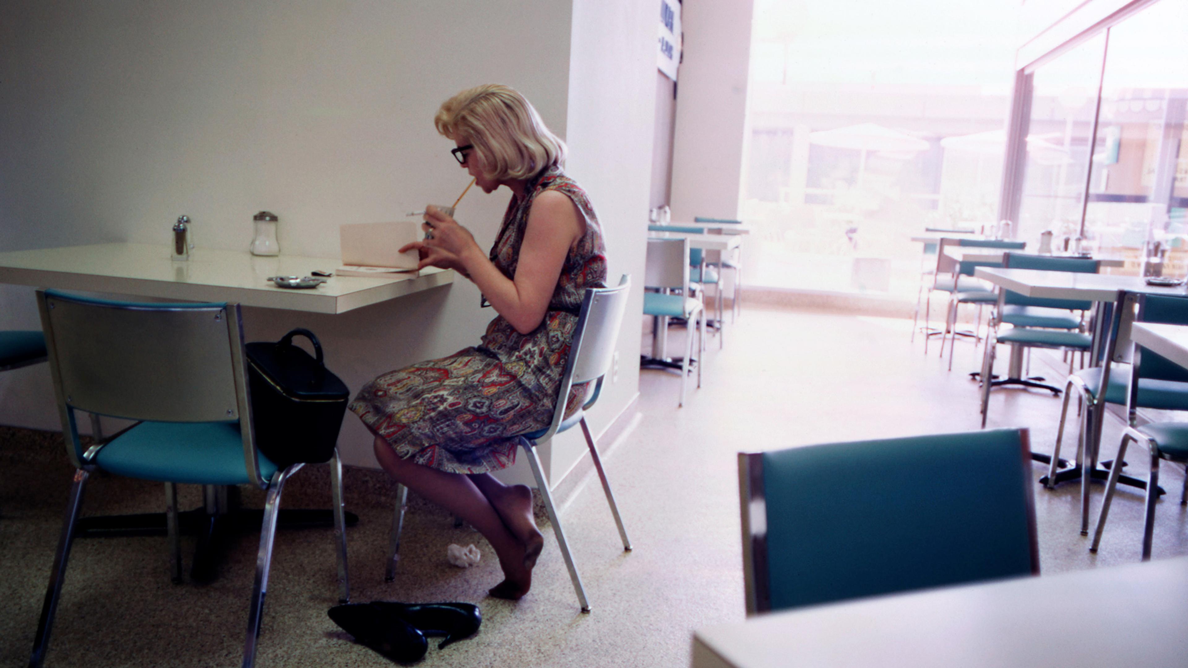 Woman seated at a cafeteria table reading a book, with her shoes kicked off.