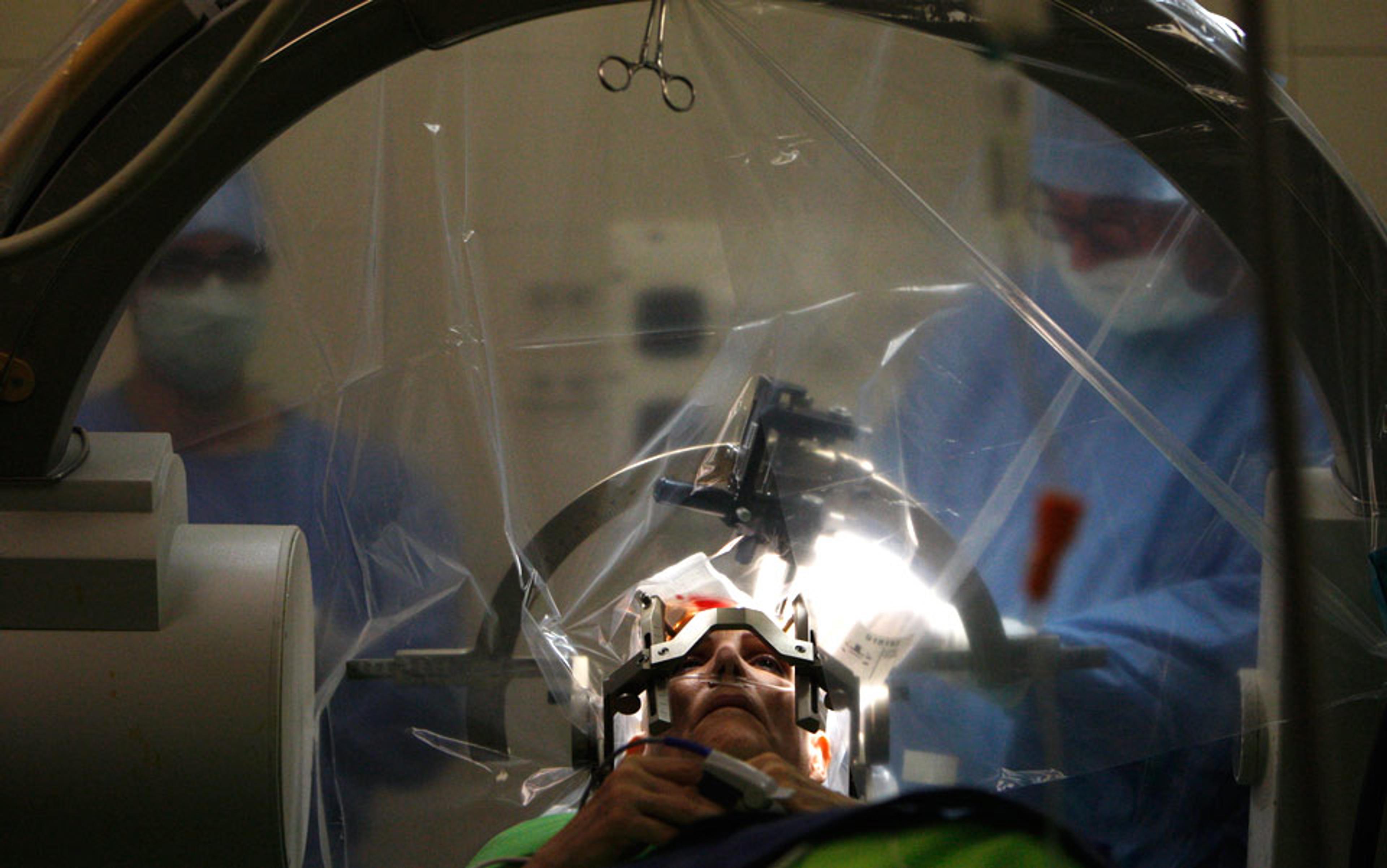 A patient in a neurosurgery procedure with two masked surgeons and medical equipment in an operating room.