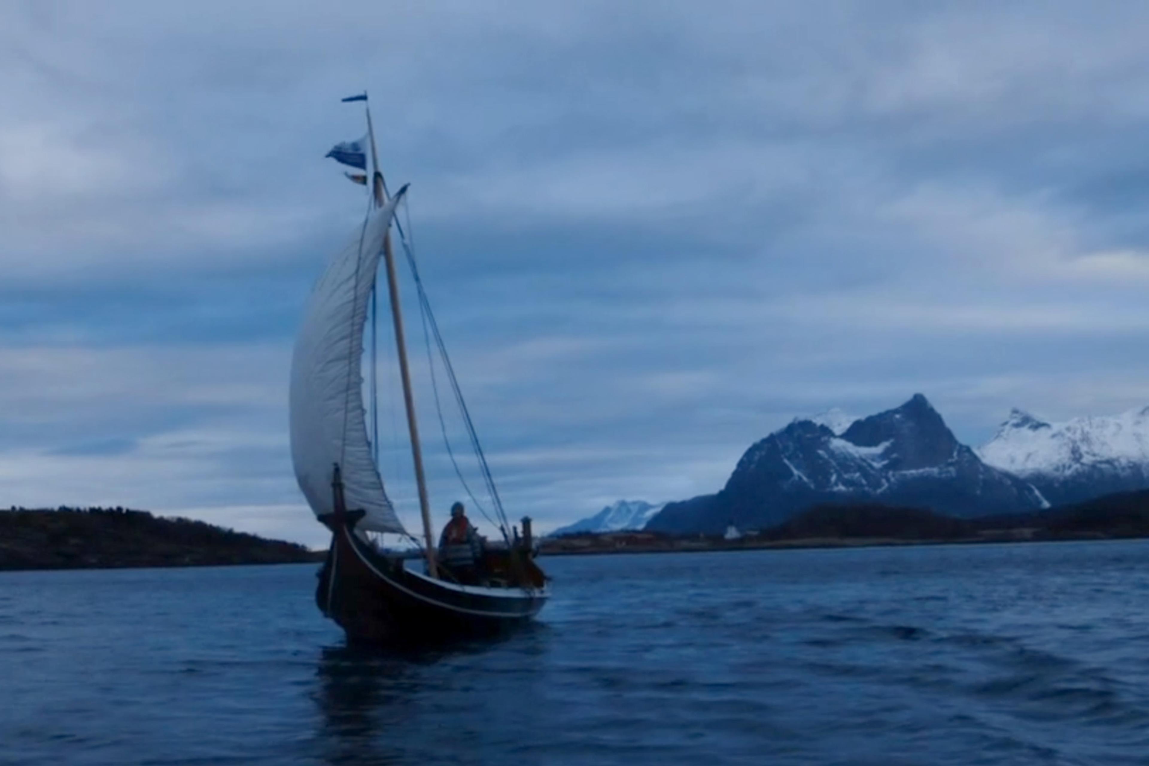 A sailboat on a calm lake at dusk with snow-capped mountains in the background under a cloudy sky.