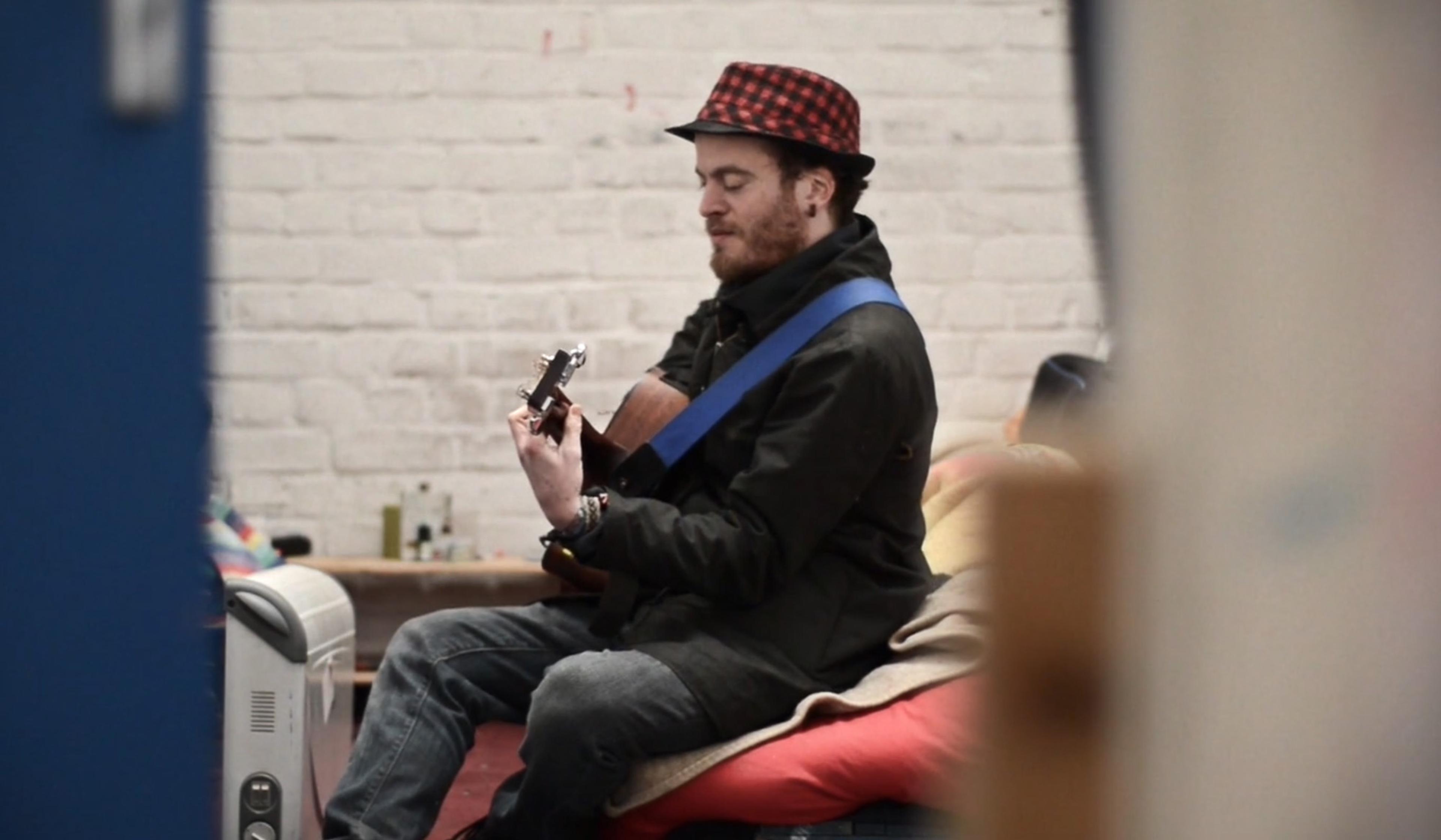Man with a beard and red checked hat plays acoustic guitar on a bed in a room with bare brick walls.