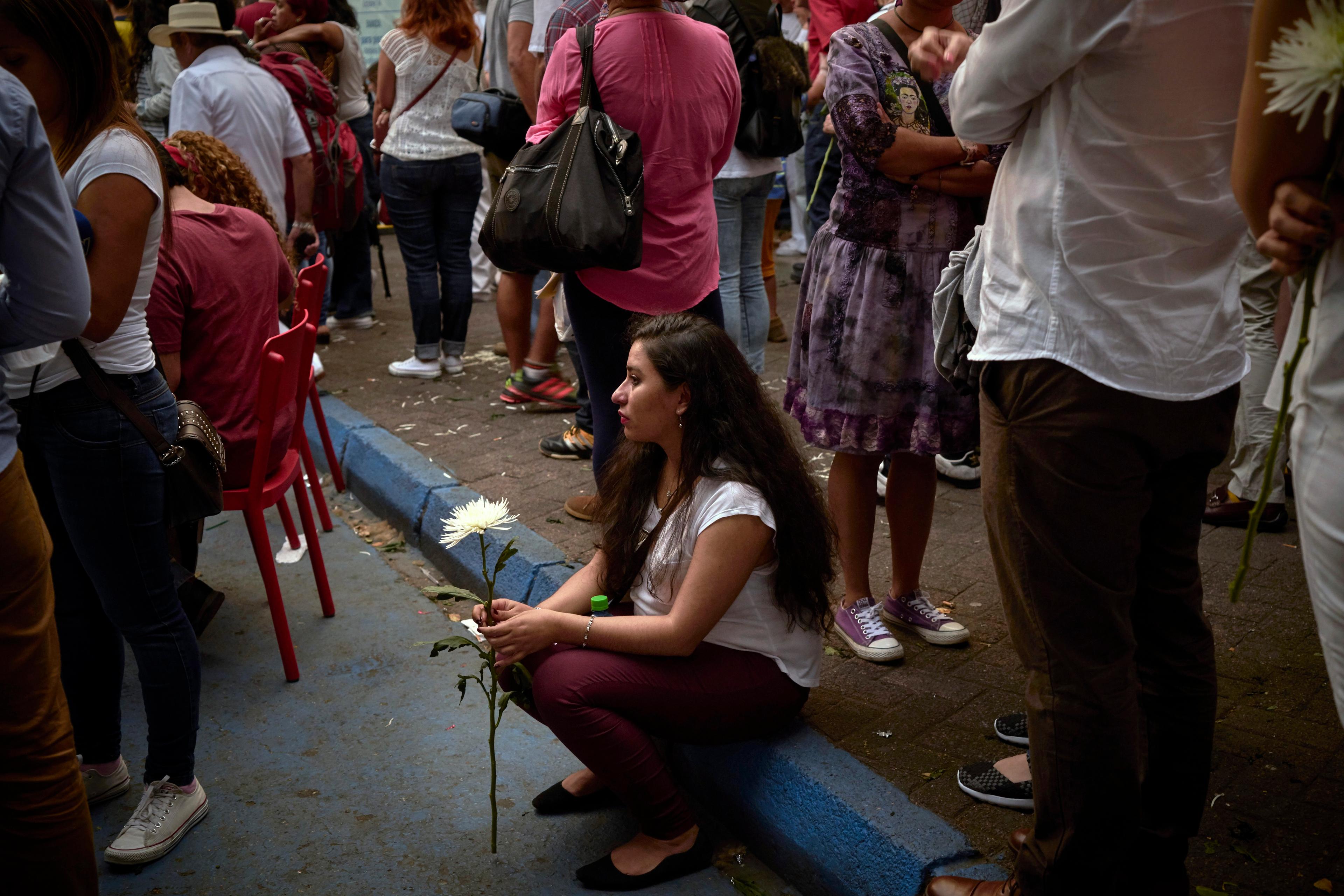 Photograph of a woman sitting on a kerb holding a white flower surrounded by people in a crowd, some standing some seated.