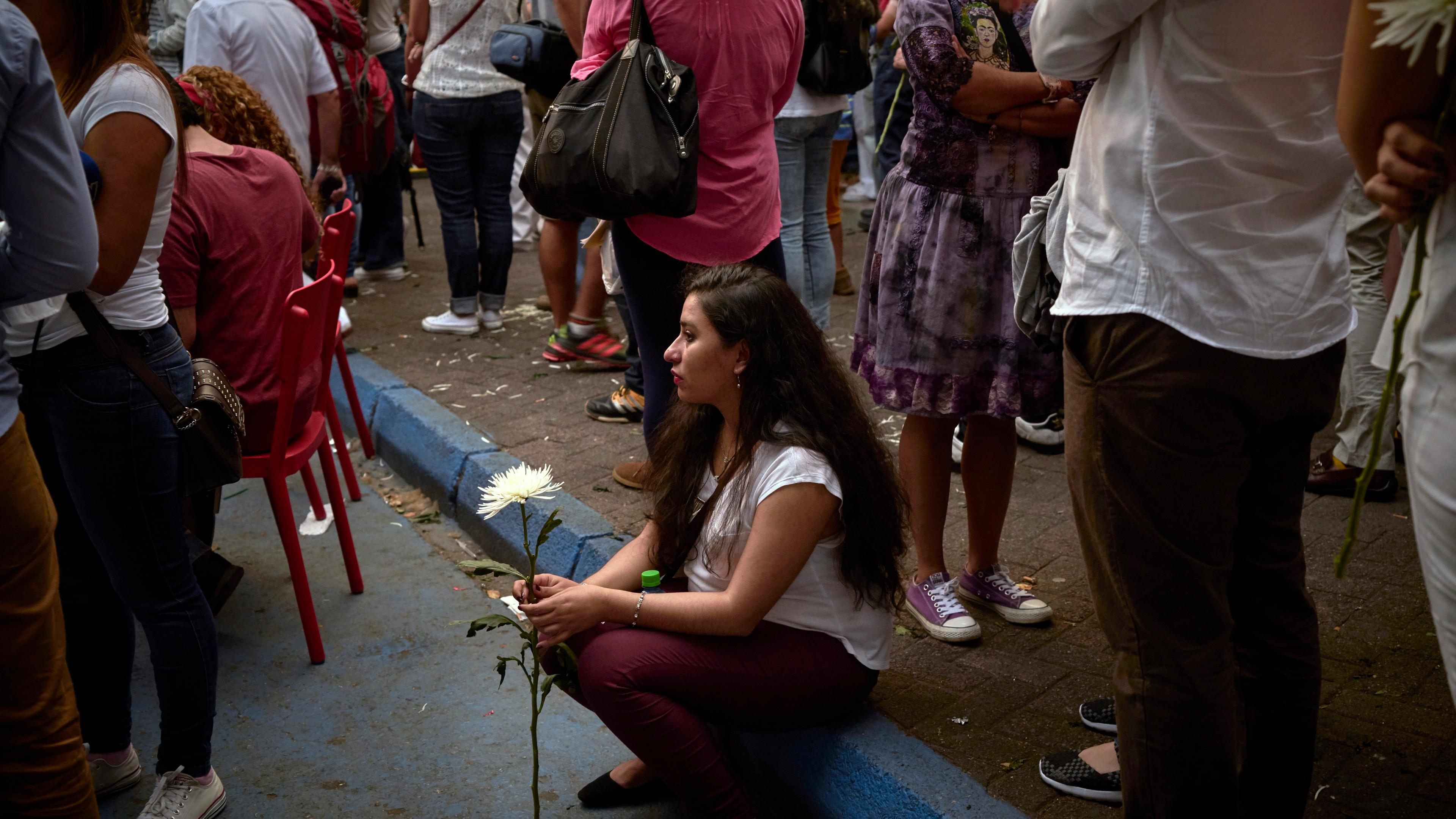 Photograph of a woman sitting on a kerb holding a white flower surrounded by people in a crowd, some standing some seated.
