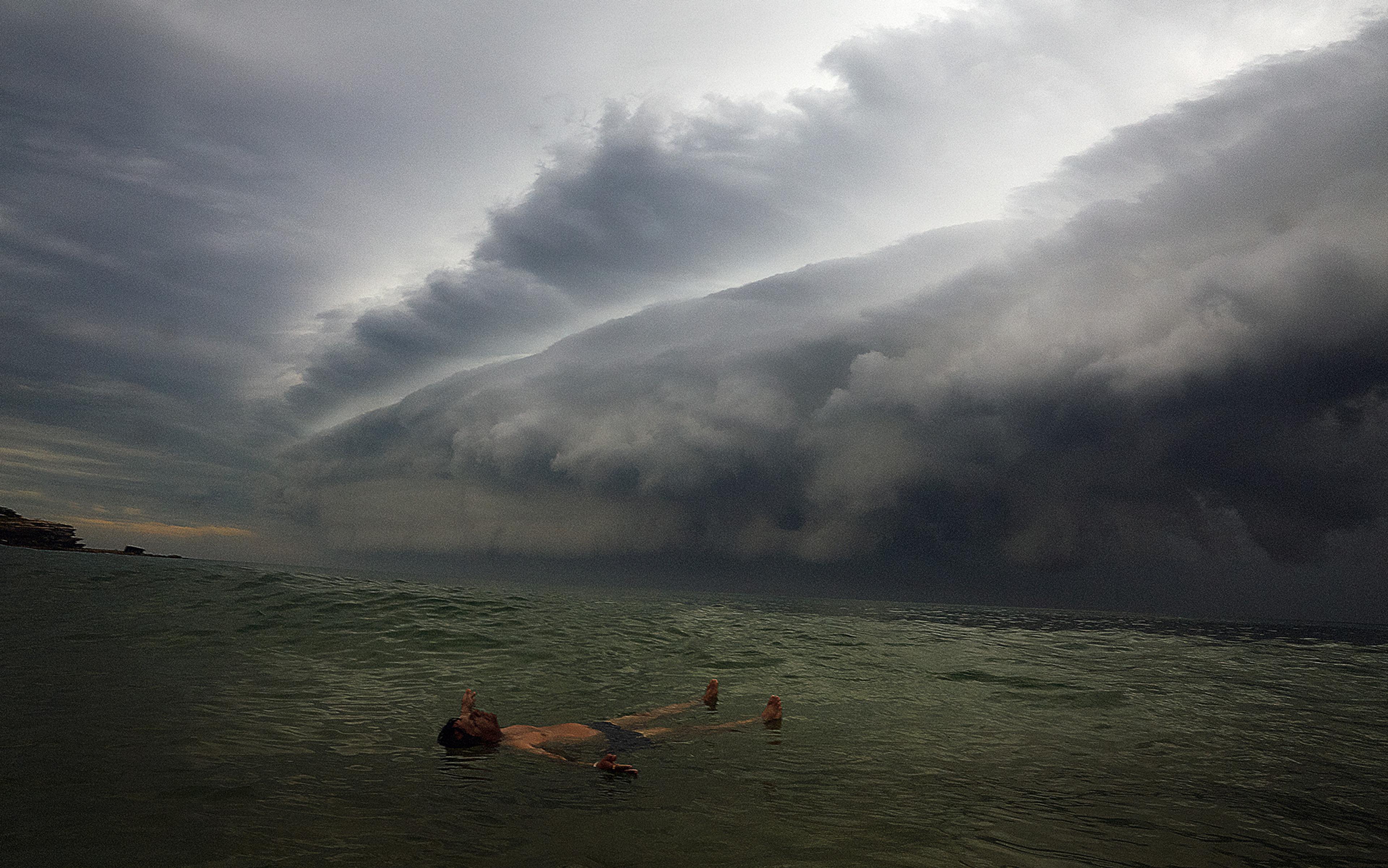 A man floating in the sea beneath a dramatic, stormy sky with large, dark clouds.