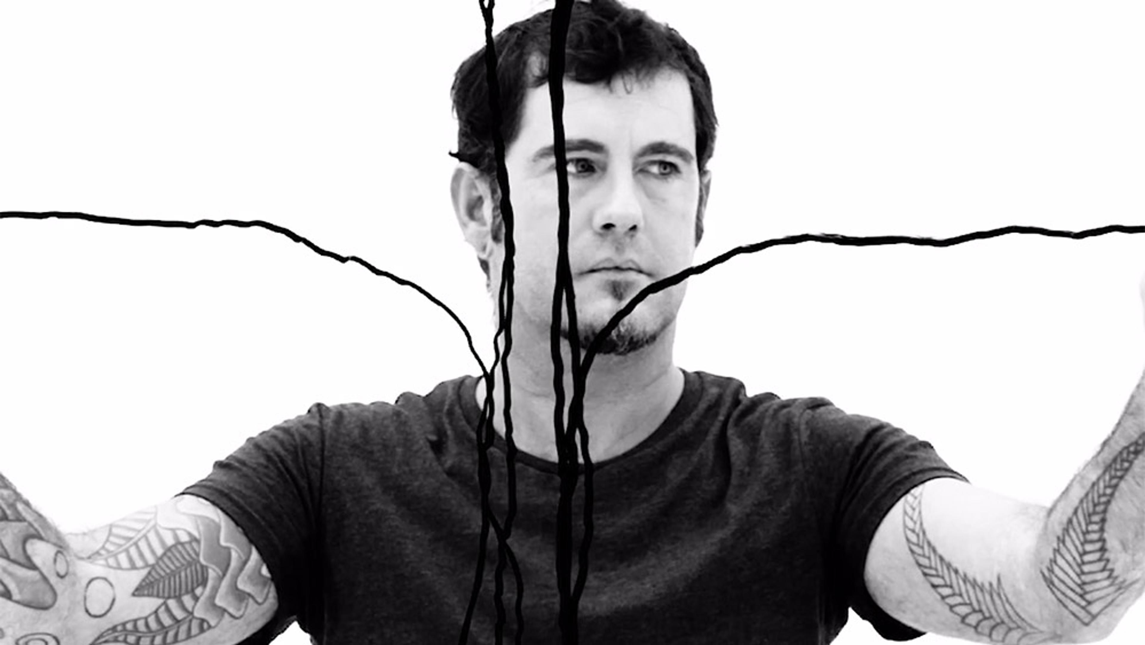 Black and white photo of a tattooed man with wires in front of him against a plain background, serious expression on his face.