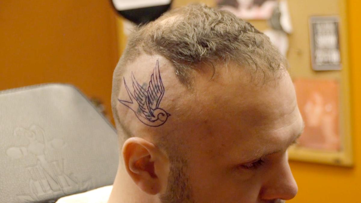 A man with a bird tattoo outlined over an older tattoo of SS insignia on his shaved head, in a tattoo parlour with orange walls.