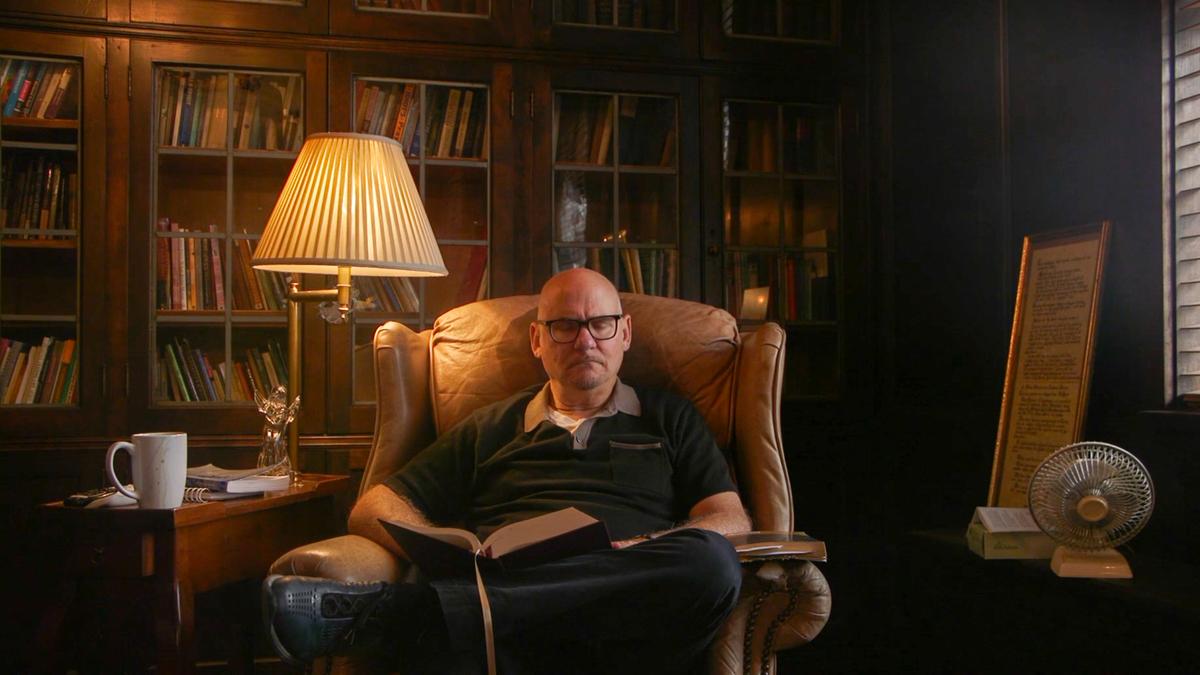 Photo of a man reading in an armchair by a lamp and bookshelf, with a side table and framed text nearby.