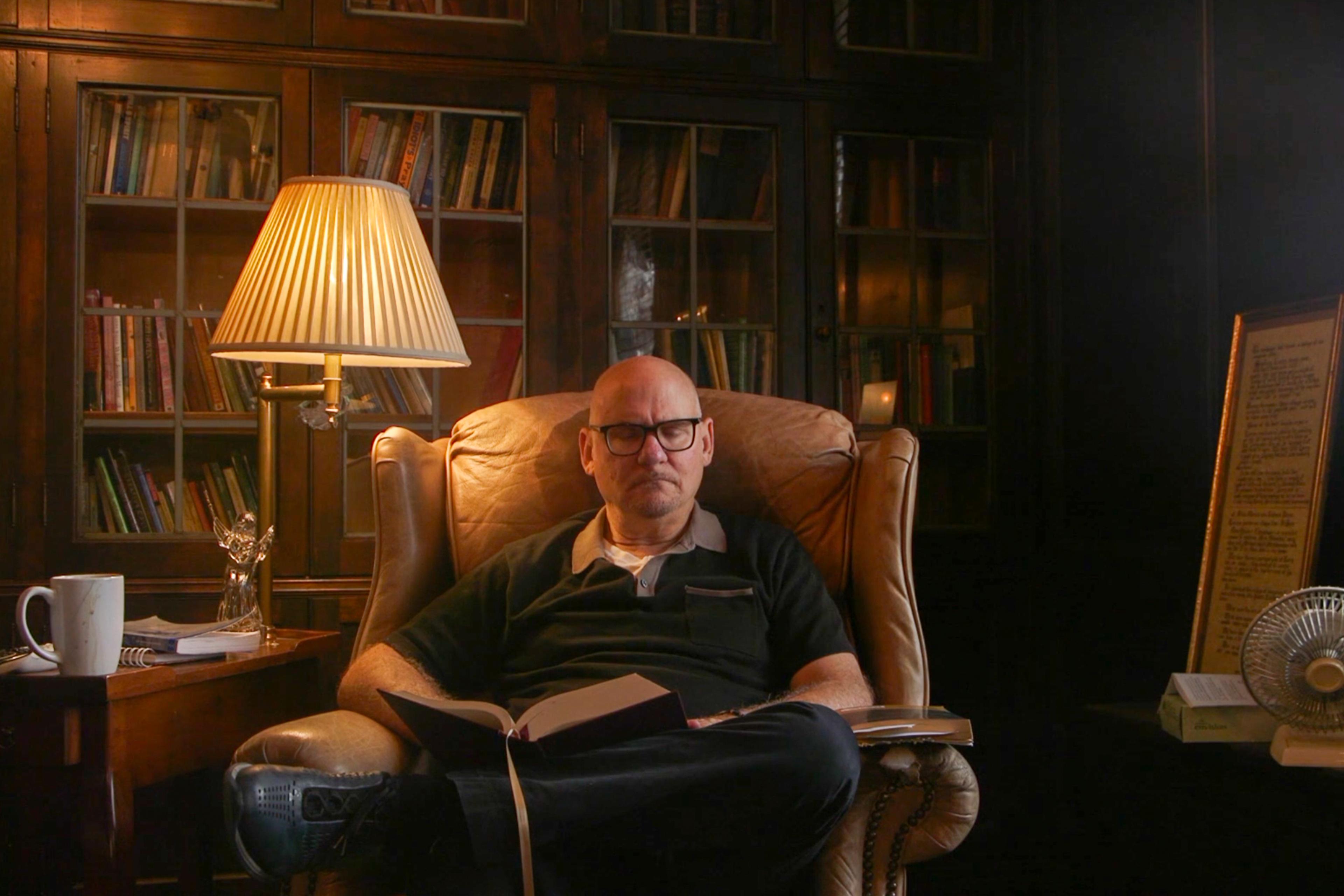 Photo of a man reading in an armchair by a lamp and bookshelf, with a side table and framed text nearby.
