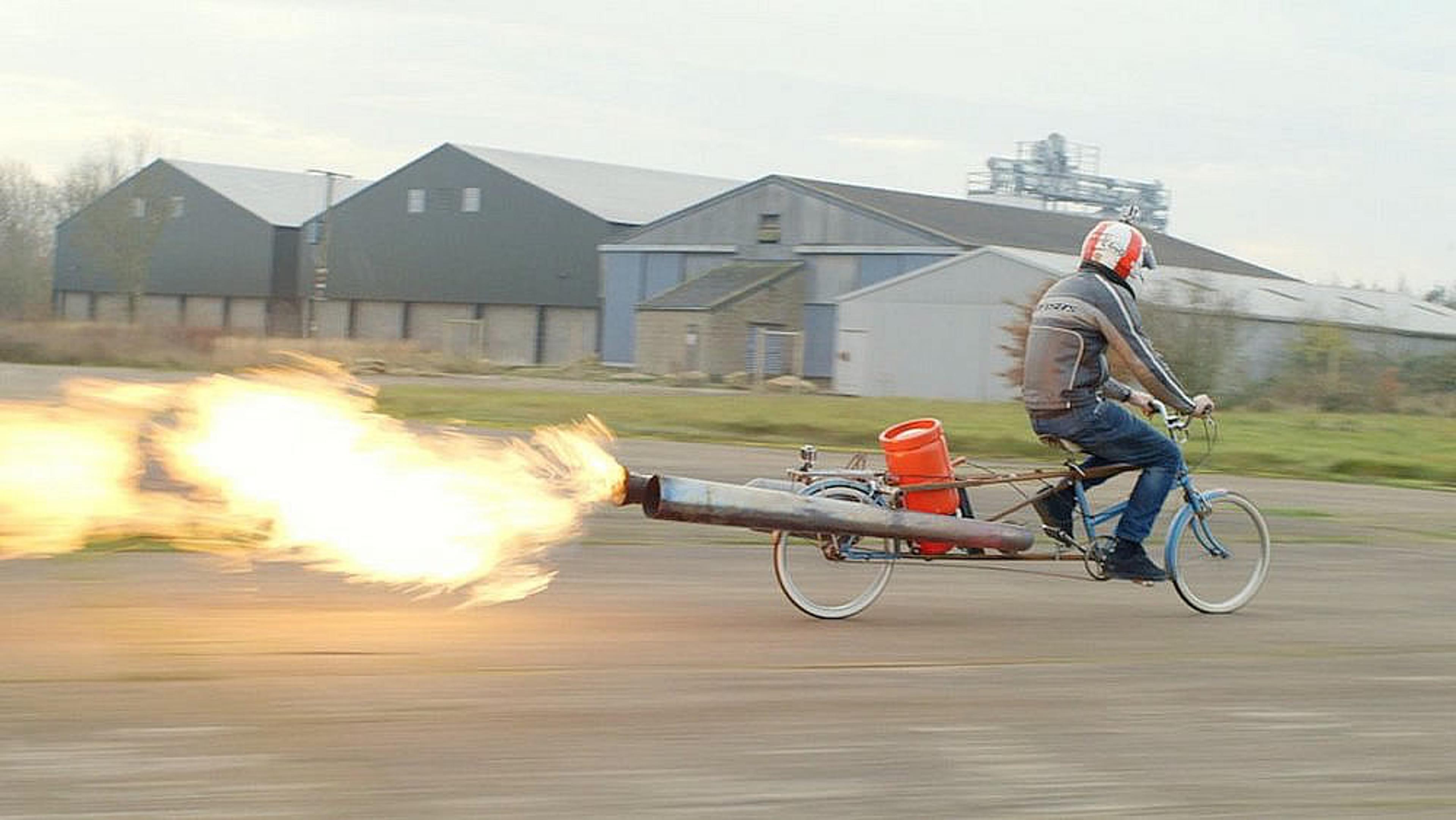 A man riding a bicycle with a large jet engine emitting flames, industrial buildings in the background.