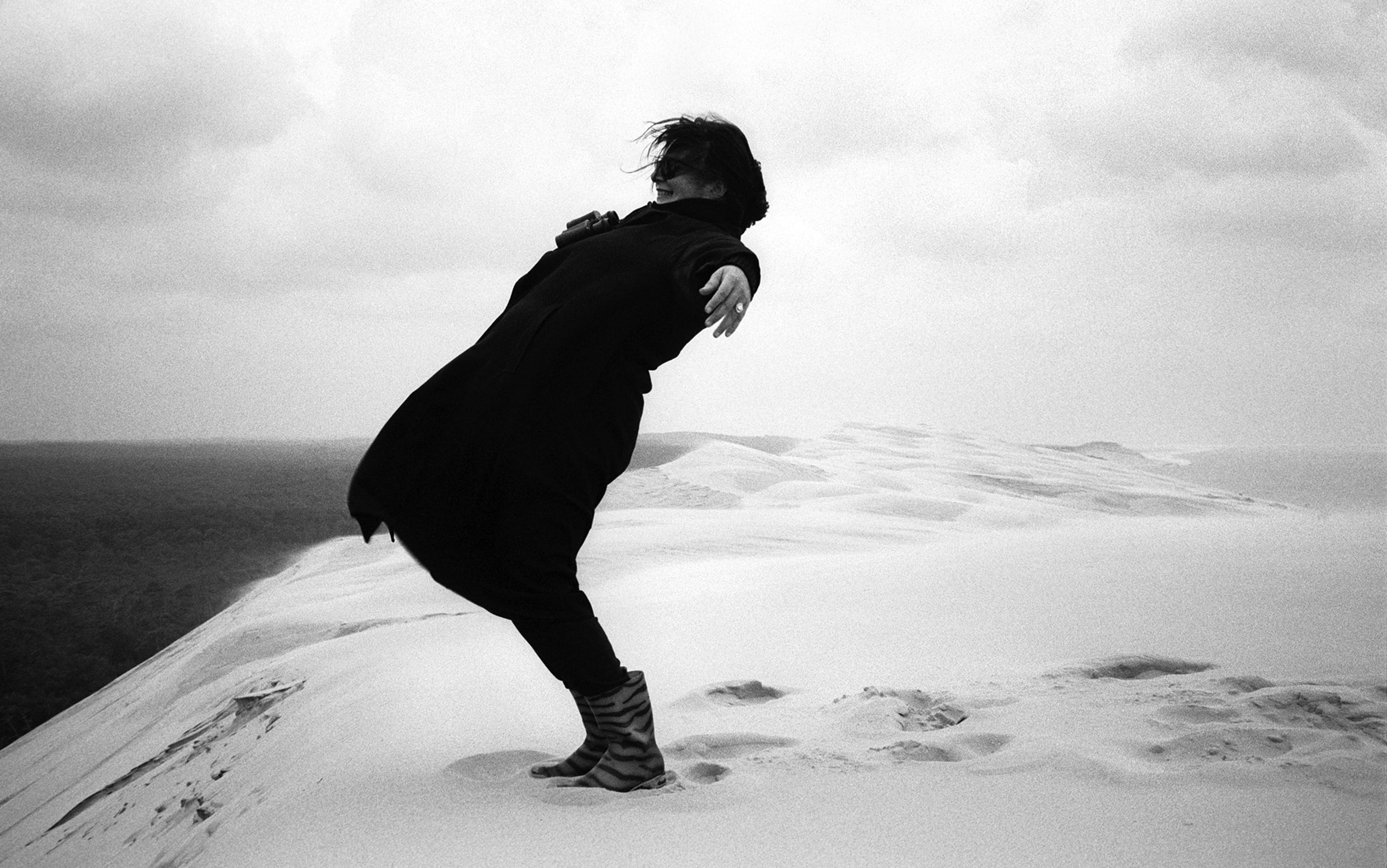 A black and white photograph shows a woman on the edge of a sand dune overlooking the sea leaning back into a strong wind