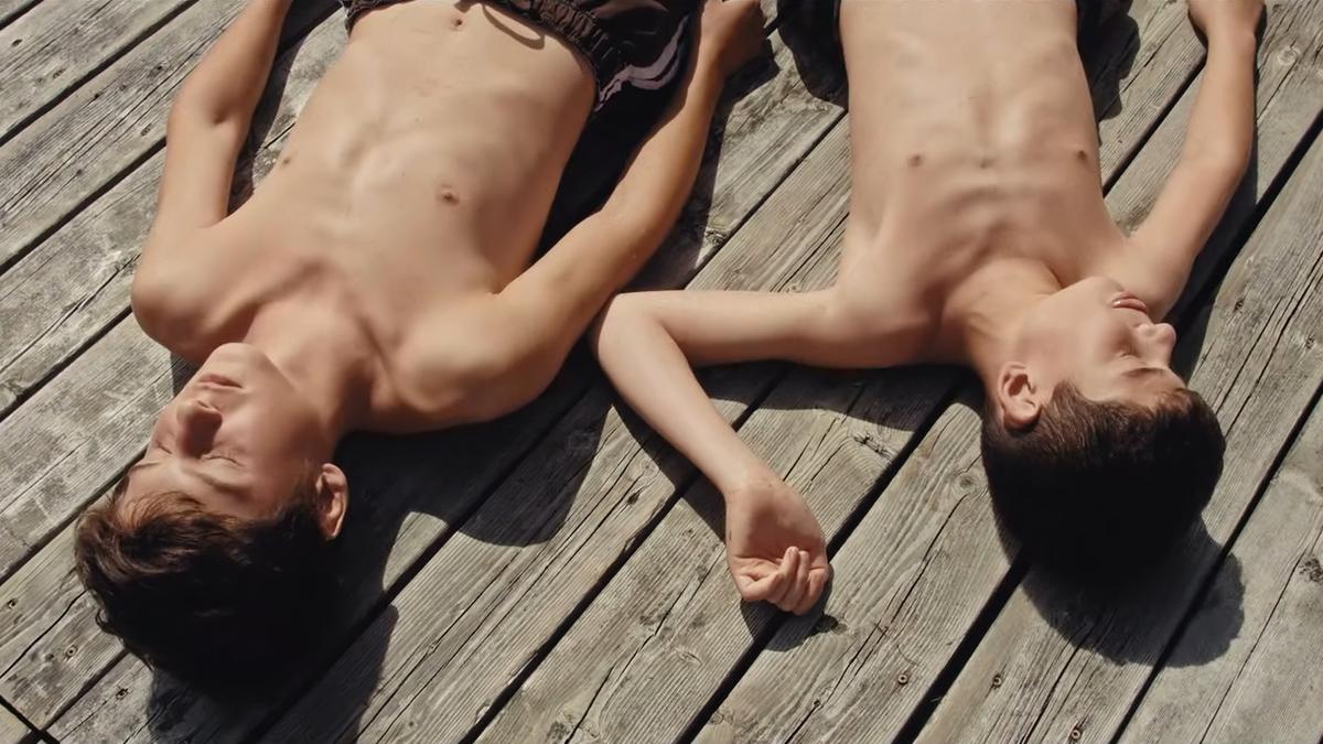 Photo of two boys in their early teens lying shirtless on a wooden deck in the sun, their eyes closed and arms resting beside them.