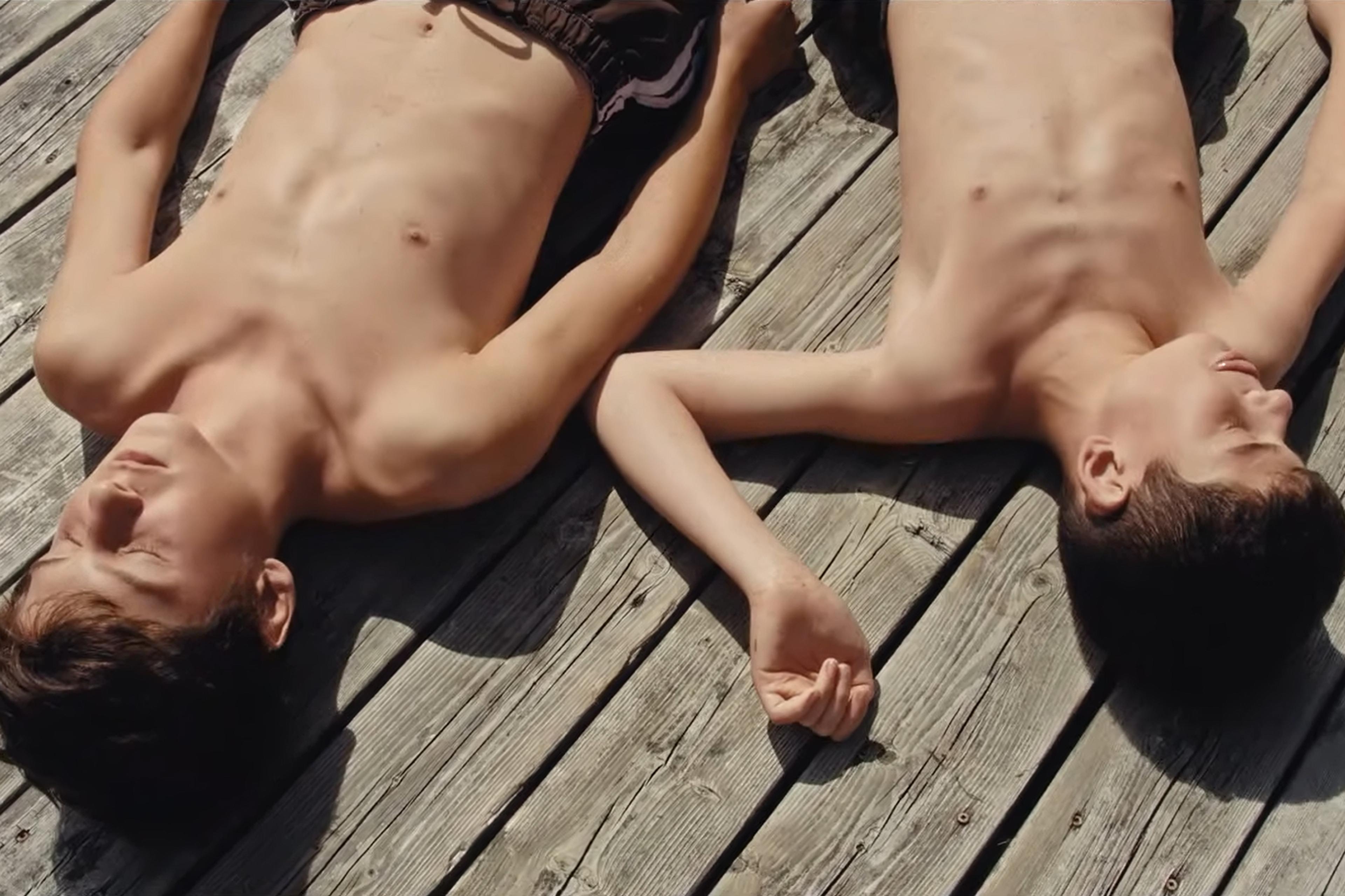 Photo of two boys in their early teens lying shirtless on a wooden deck in the sun, their eyes closed and arms resting beside them.