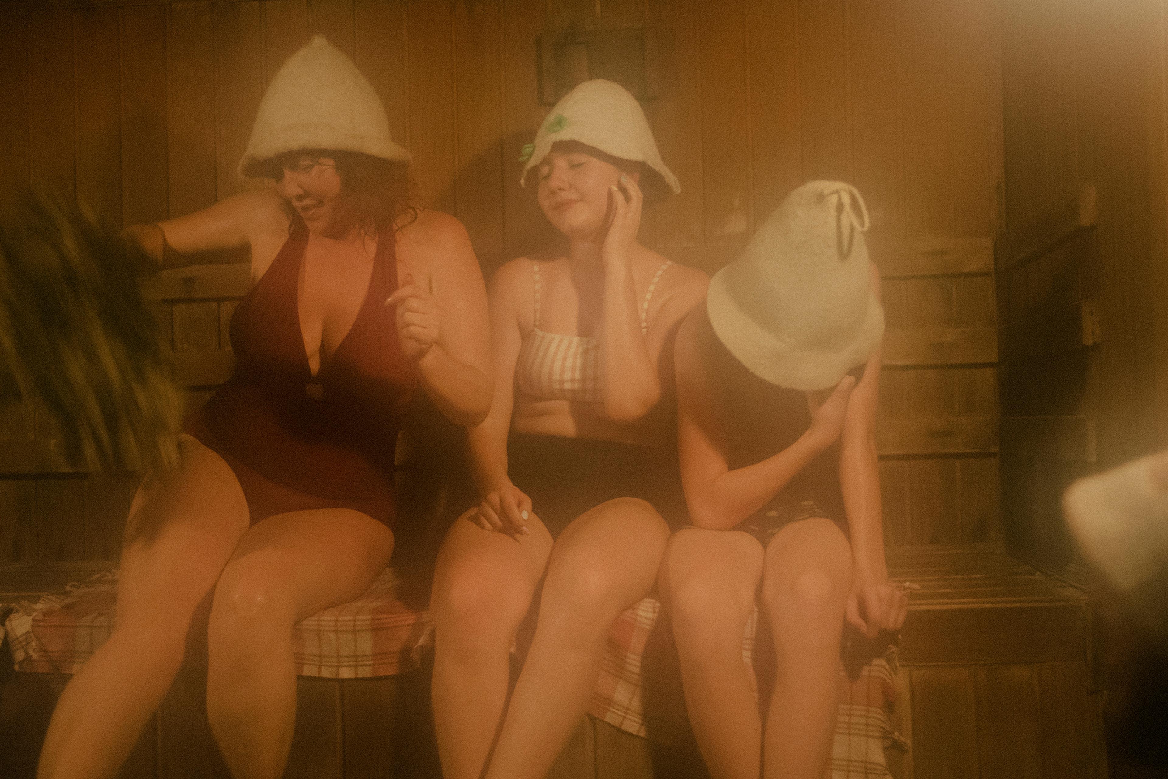 Three people in swimwear wearing felt hats sitting in a sauna. One holds branches, the atmosphere is warm and misty.