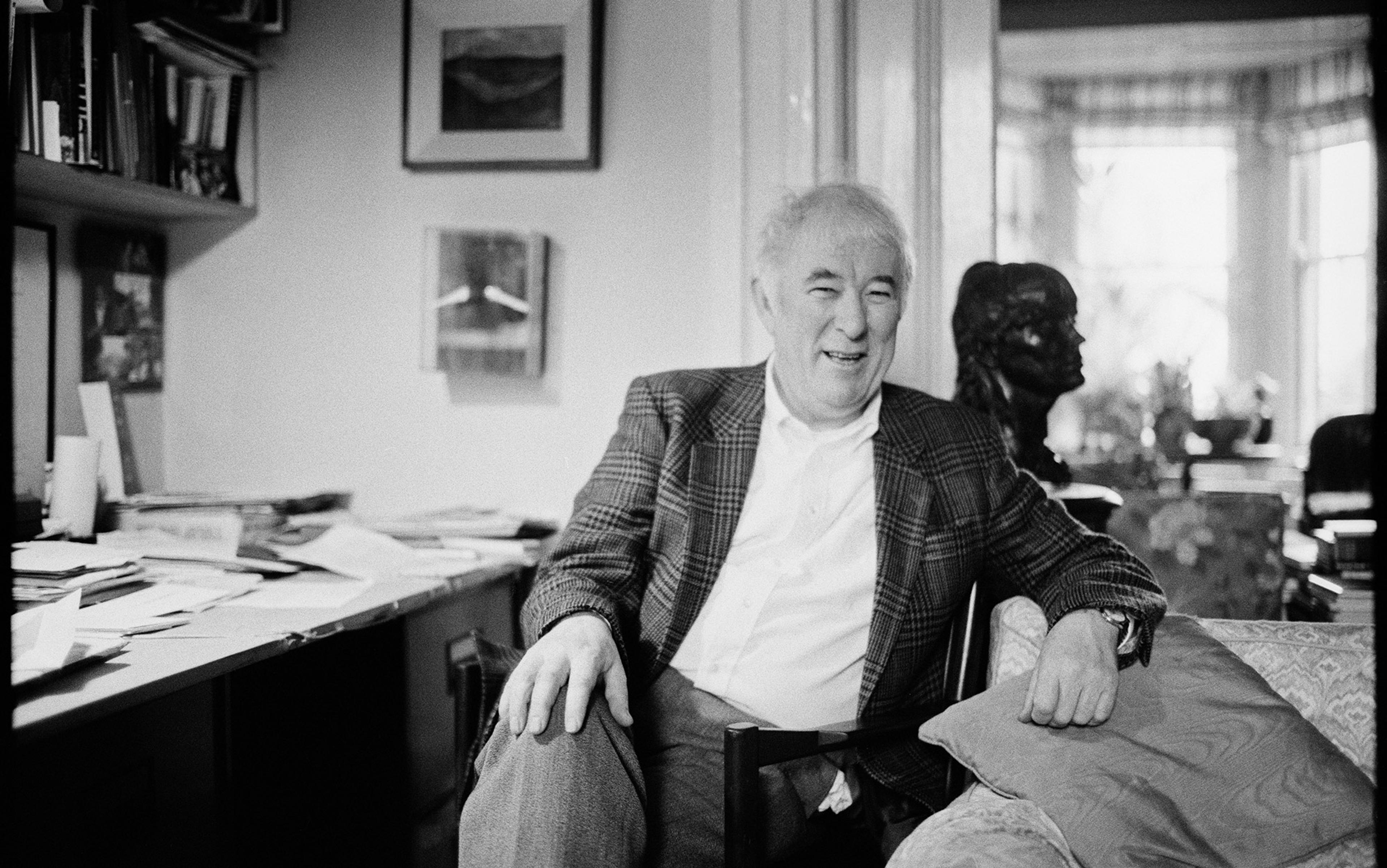 A black and white photo of the poet Seamus Heaney sitting in a cosy, cluttered study, smiling next to a desk filled with papers and books.