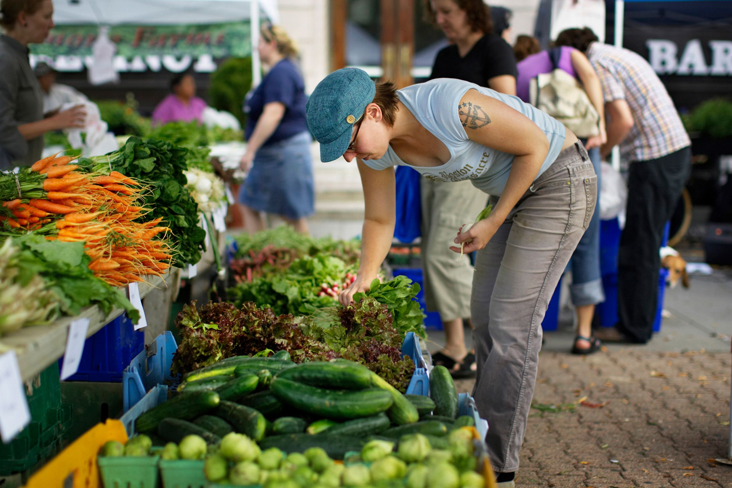 A woman selecting vegetables at an outdoor market stall with assorted greens and carrots available for sale.