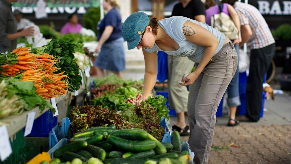 A woman selecting vegetables at an outdoor market stall with assorted greens and carrots available for sale.