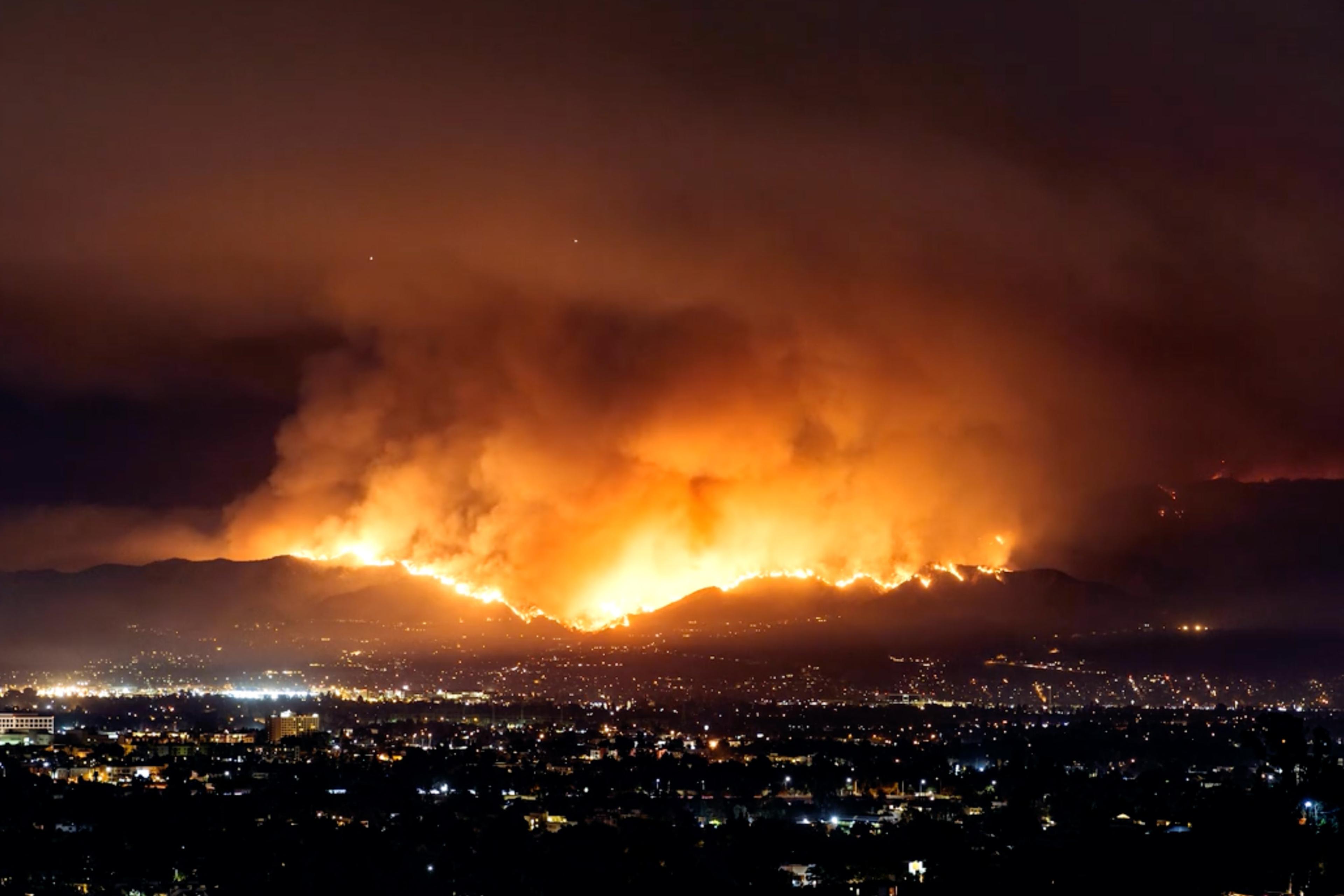 Video-loop of a night-time cityscape with a large wildfire blazing on distant hills under a smoky sky.