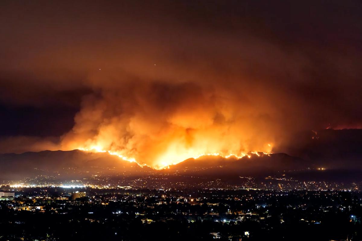 Video-loop of a night-time cityscape with a large wildfire blazing on distant hills under a smoky sky.