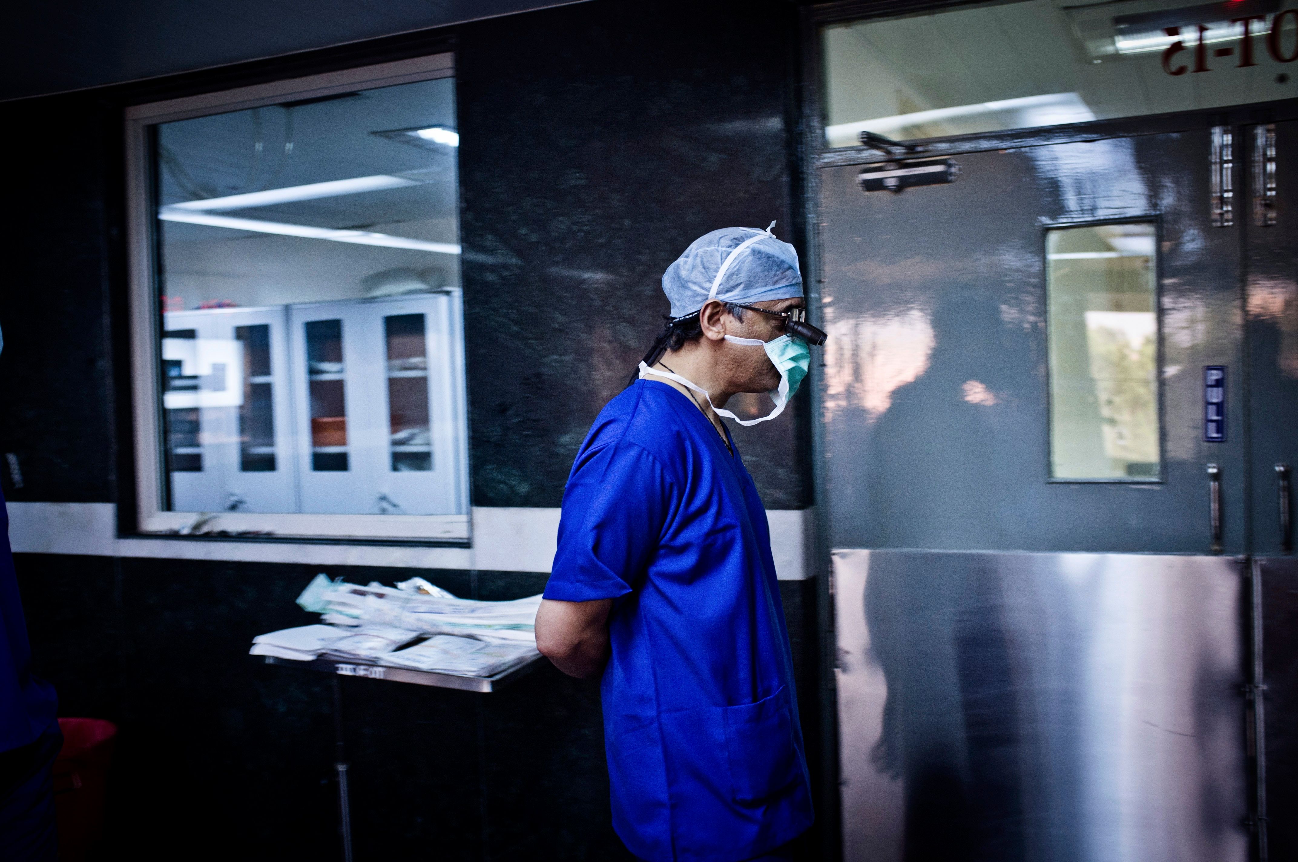 Photo of a surgeon in blue scrubs and mask walking in a hospital hallway near a surgical prep area.