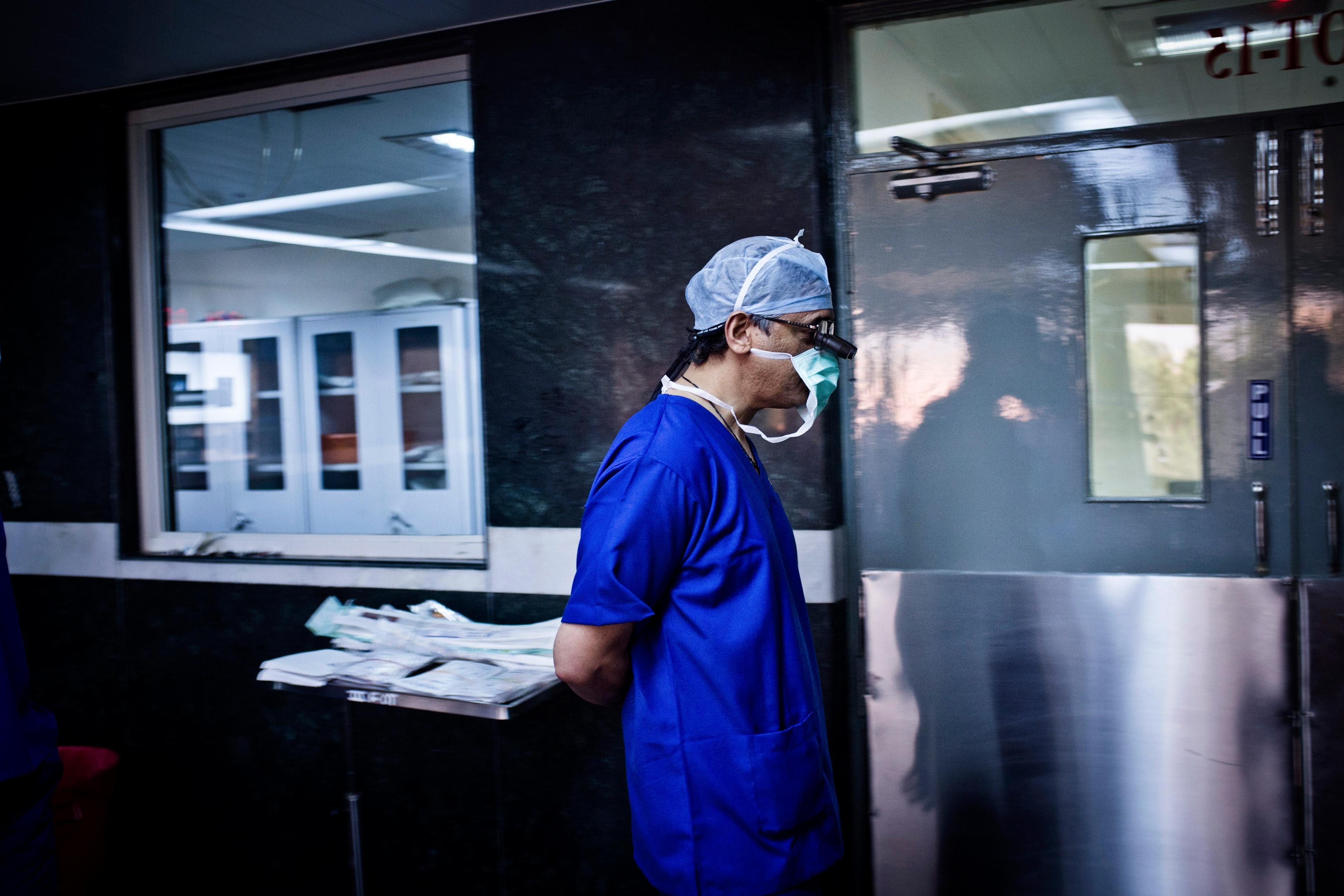 Photo of a surgeon in blue scrubs and mask walking in a hospital hallway near a surgical prep area.