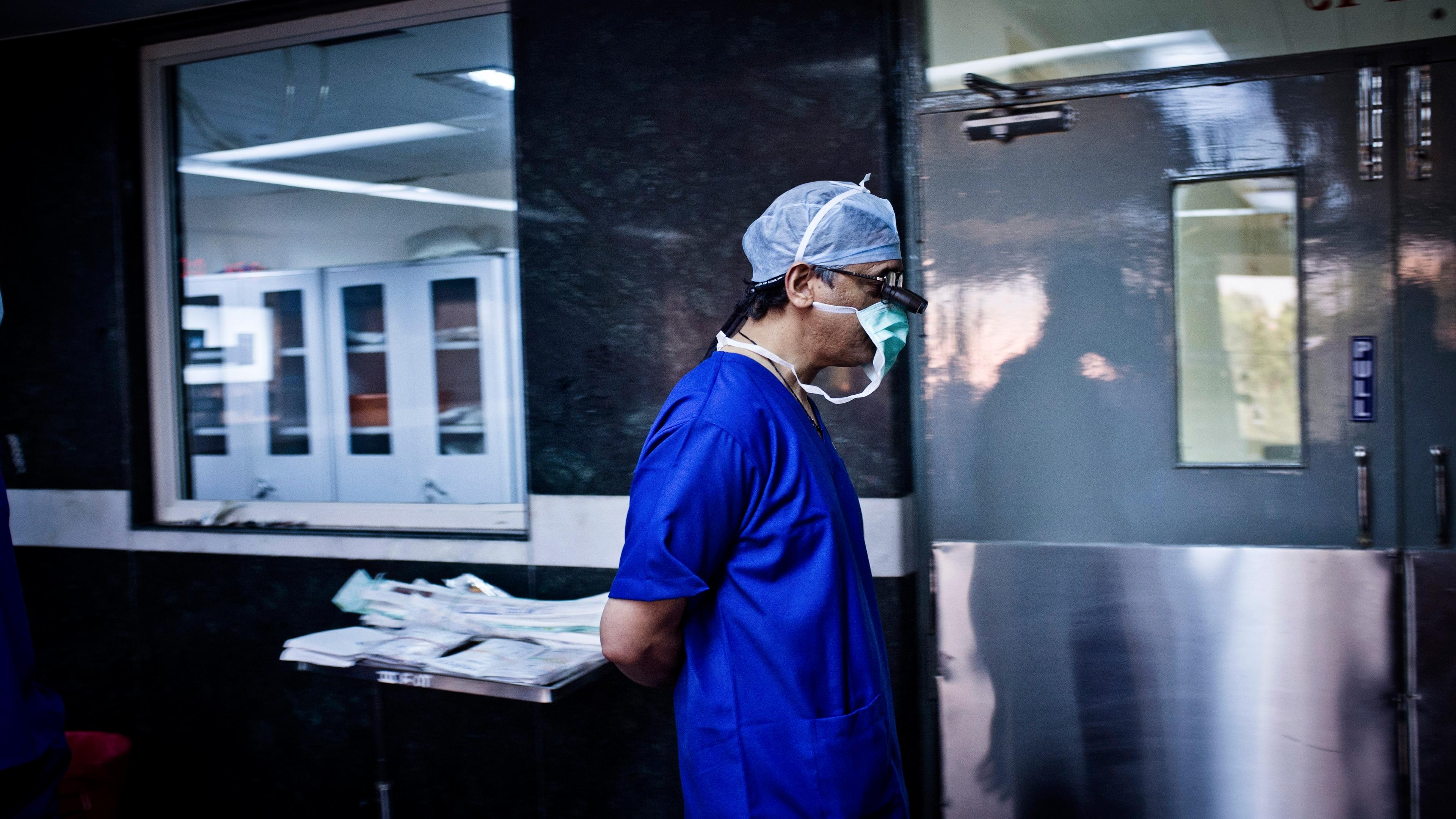 Photo of a surgeon in blue scrubs and mask walking in a hospital hallway near a surgical prep area.