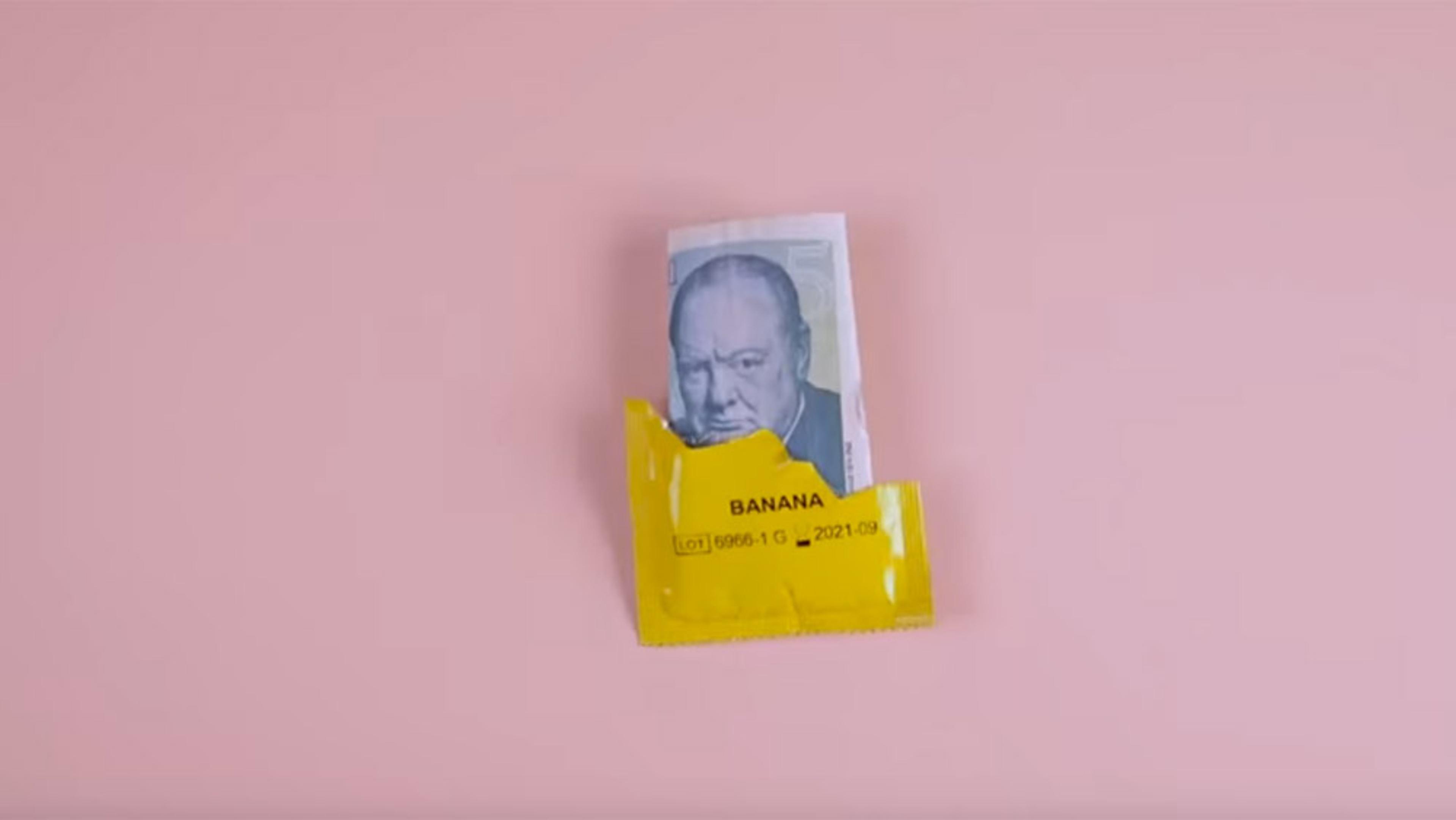 A folded sterling banknote, partially wrapped in yellow packaging labelled ‘BANANA’ against a pink background.