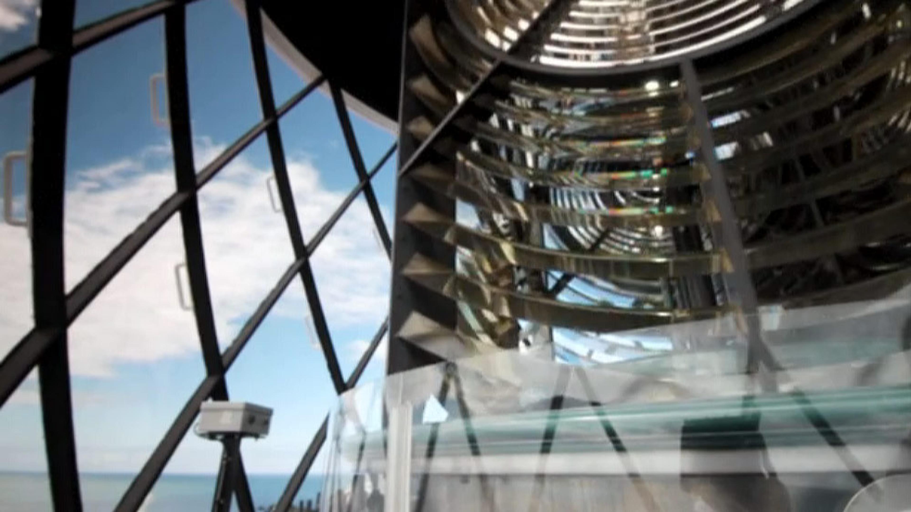 A large lens, likely from a lighthouse, with a view of the sea and sky through window panes on the left side.