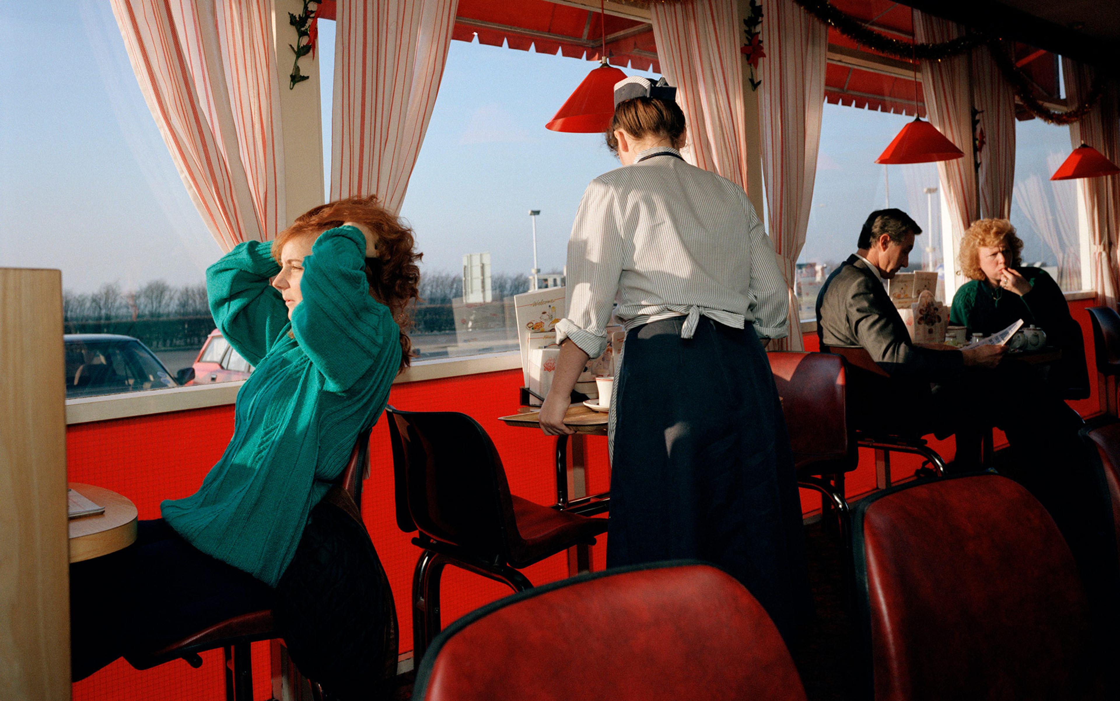 People seated in a diner with red accents, a waitress serving, and a woman in a green sweater sitting by the window adjusting her hair.