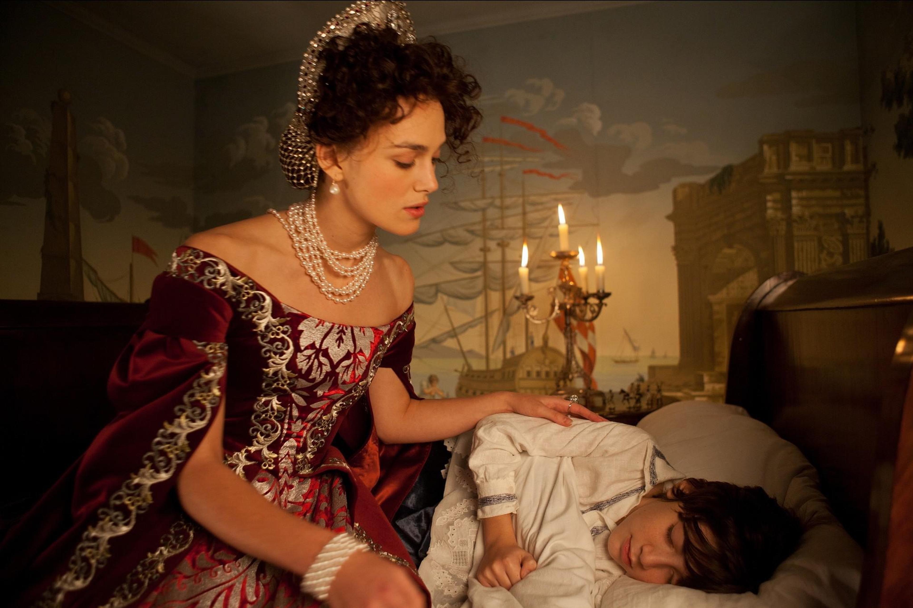 A photo of a woman in historical attire watching over a sleeping child by candlelight, with a mural of ships in the background.